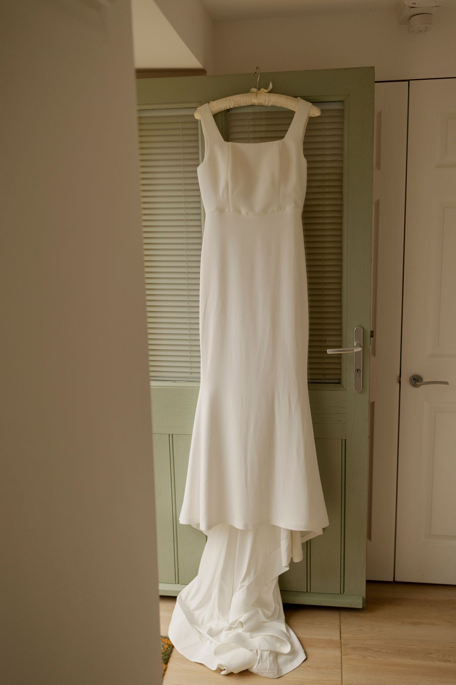 A simple white floor-length dress hangs on display in various angles against a neutral wall with blinds.