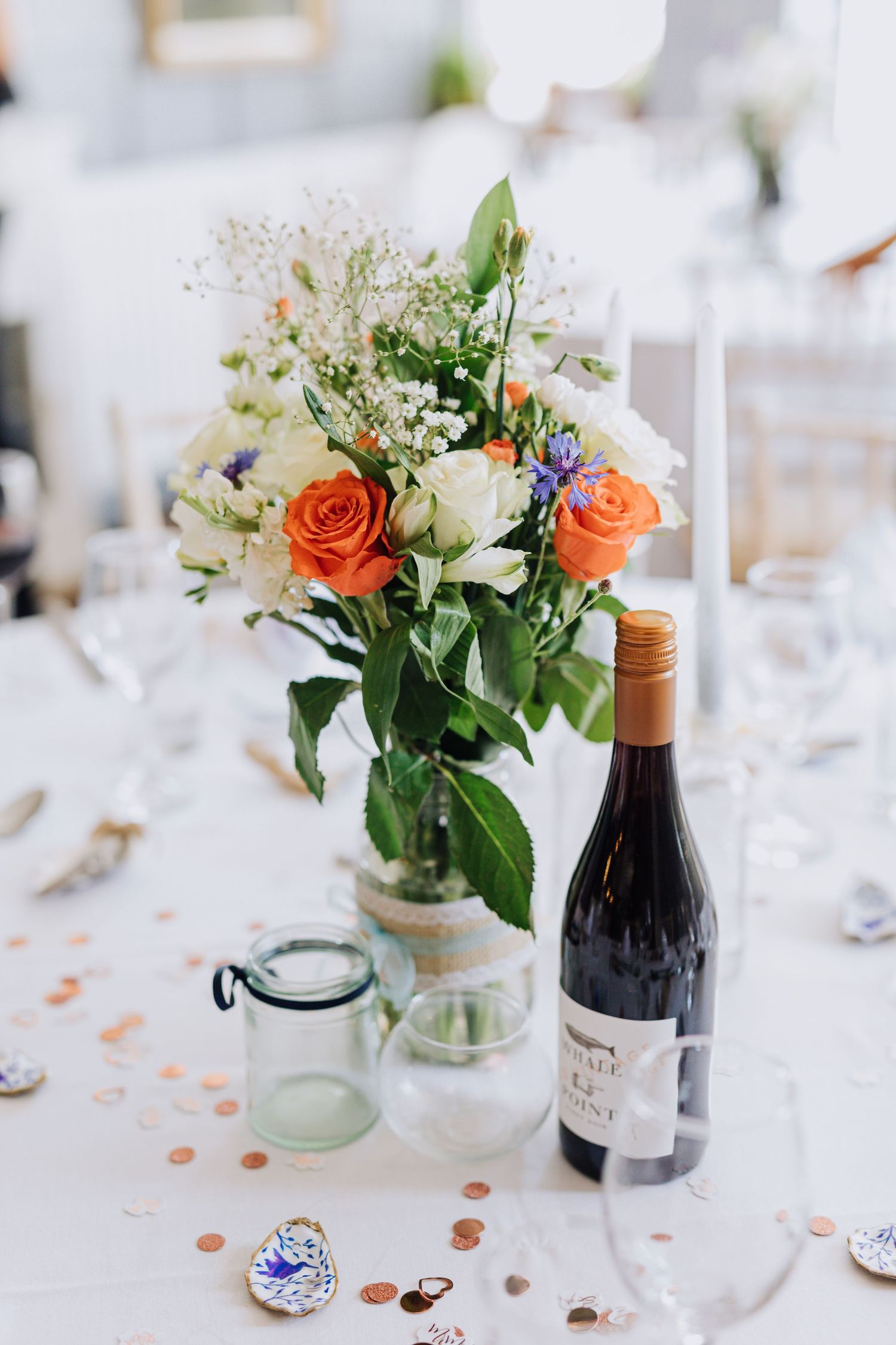 Elegant wedding table centerpiece with orange roses and white flowers next to a wine bottle on a white tablecloth.
