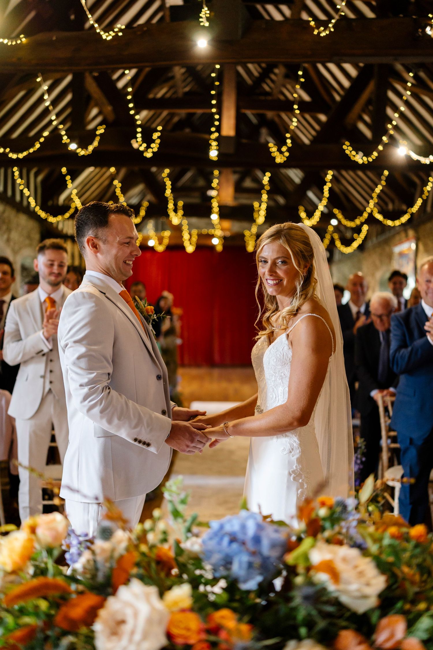 A wedding ceremony under wooden beams decorated with string lights and floral arrangements.