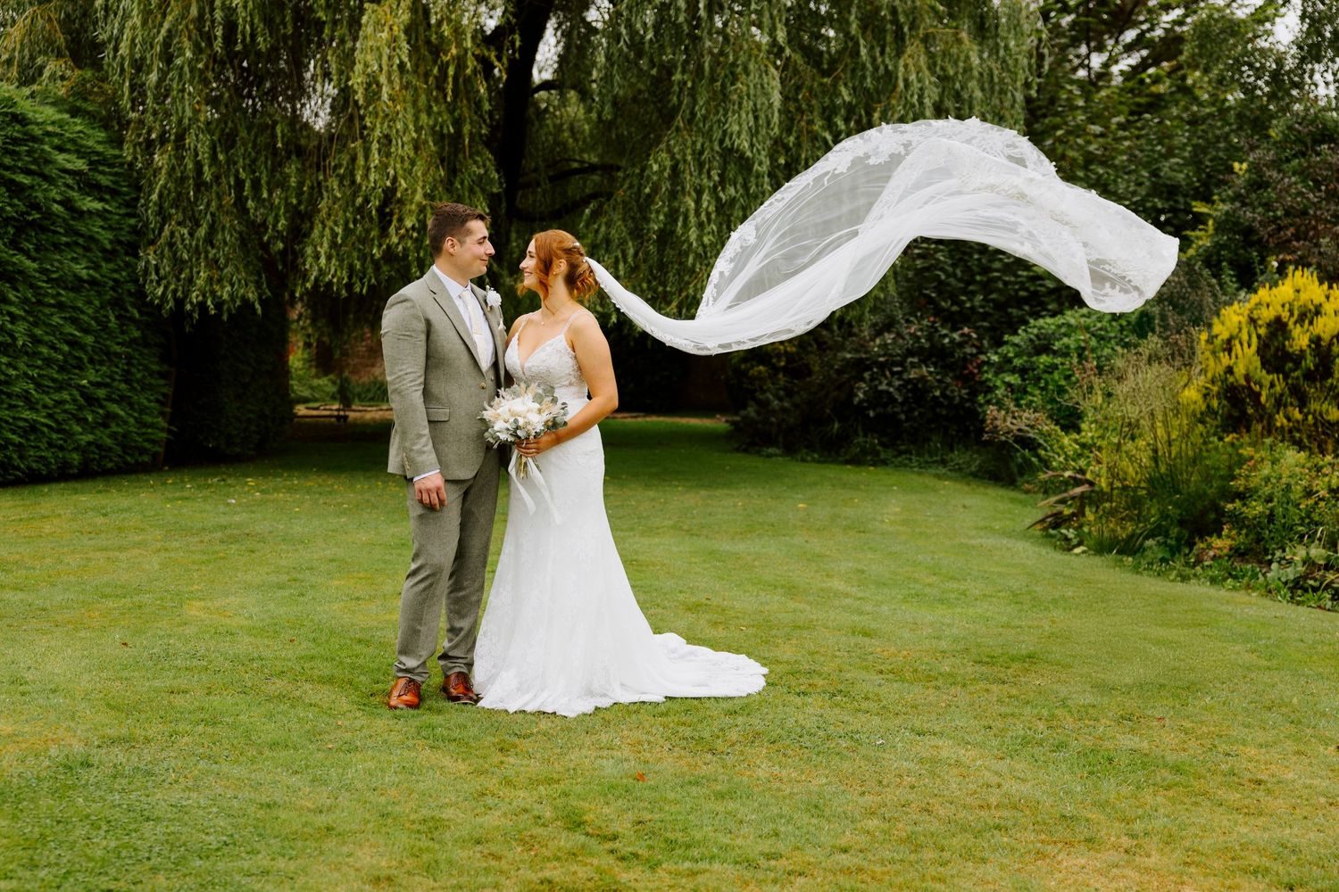 A wedding couple poses on a green lawn as the bride's veil flows dramatically in the wind against garden scenery.