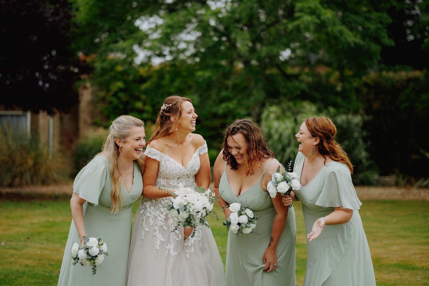 A bride in a lace gown stands with bridesmaids in mint green dresses holding white bouquets.