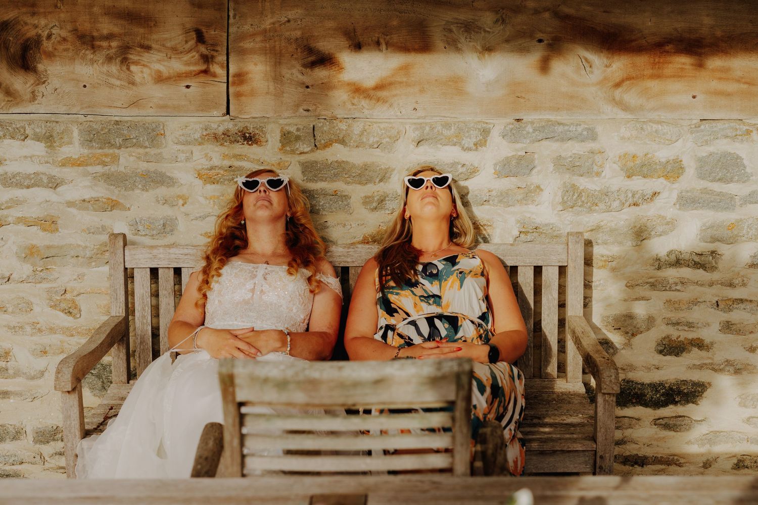 Two people relaxing on a rustic wooden bench against a stone wall.