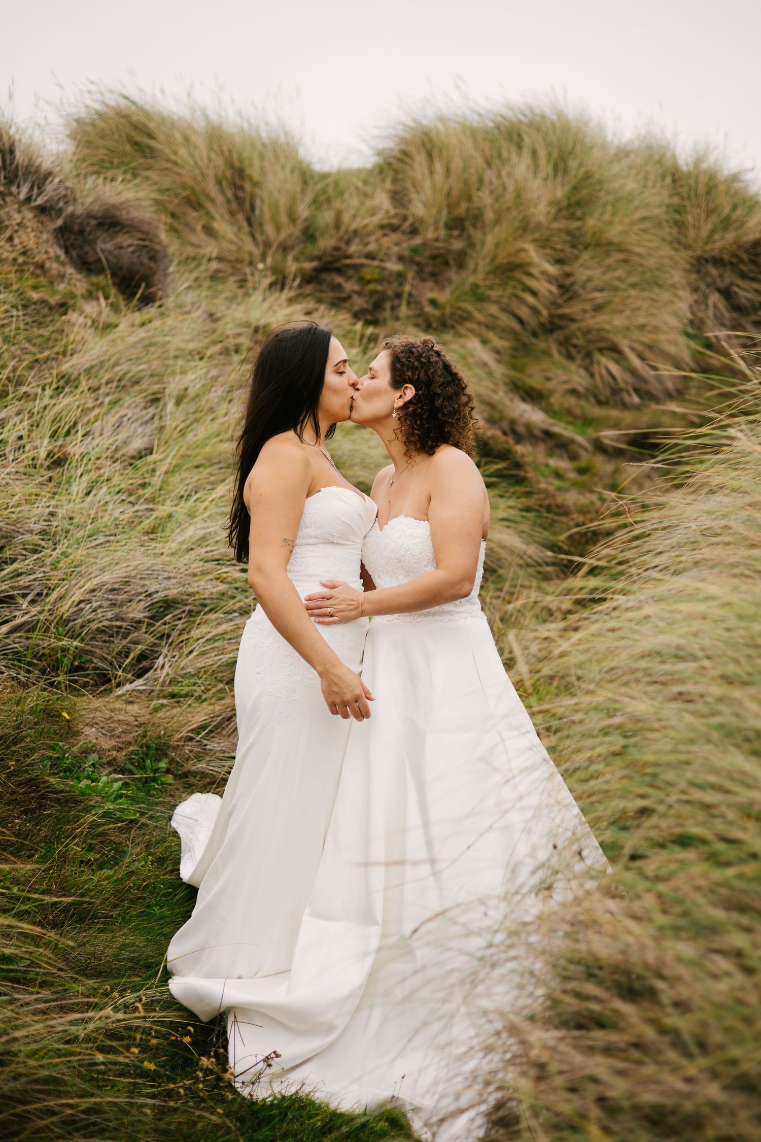 A loving moment between two brides among windswept coastal grass.