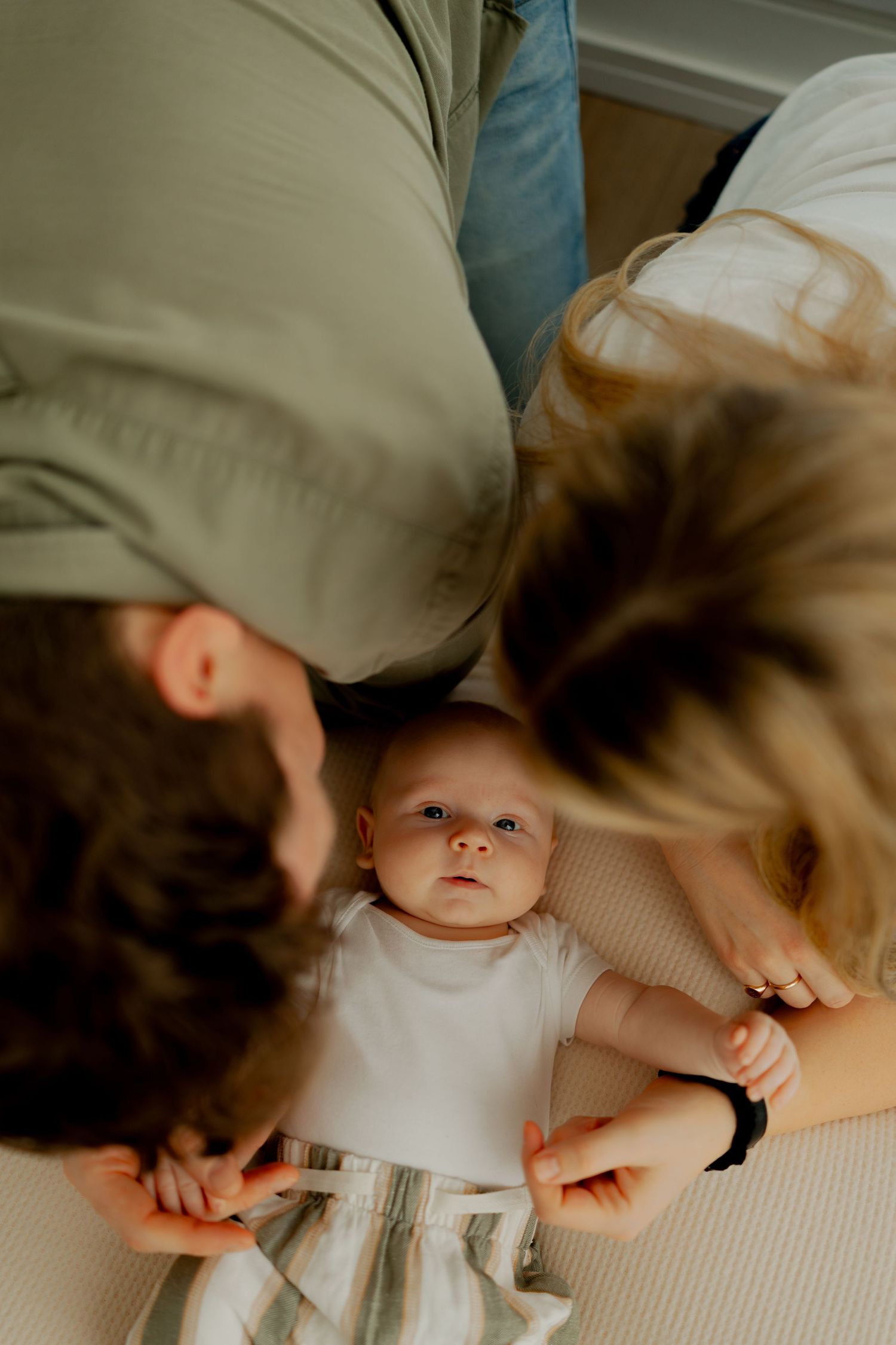A baby in a white onesie lies between two adults on a bed, looking up at the camera.