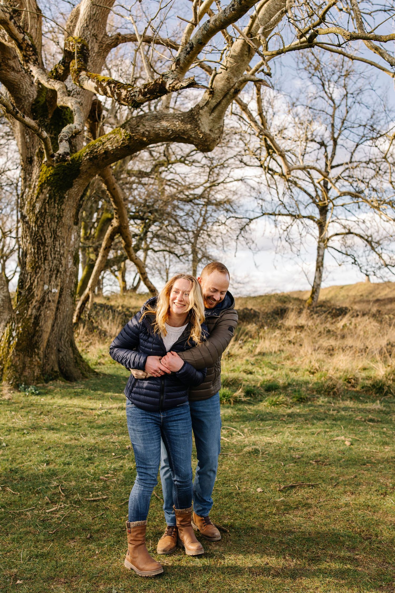 Couple embracing under large tree branches in nature setting during golden hour.