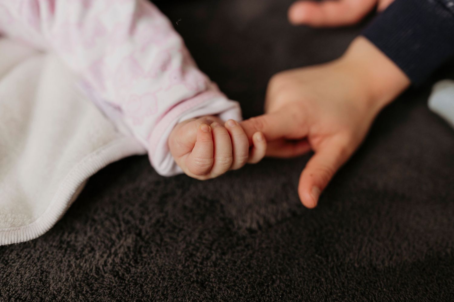 Close-up of baby's tiny hand touching an adult's palm on dark textured fabric.