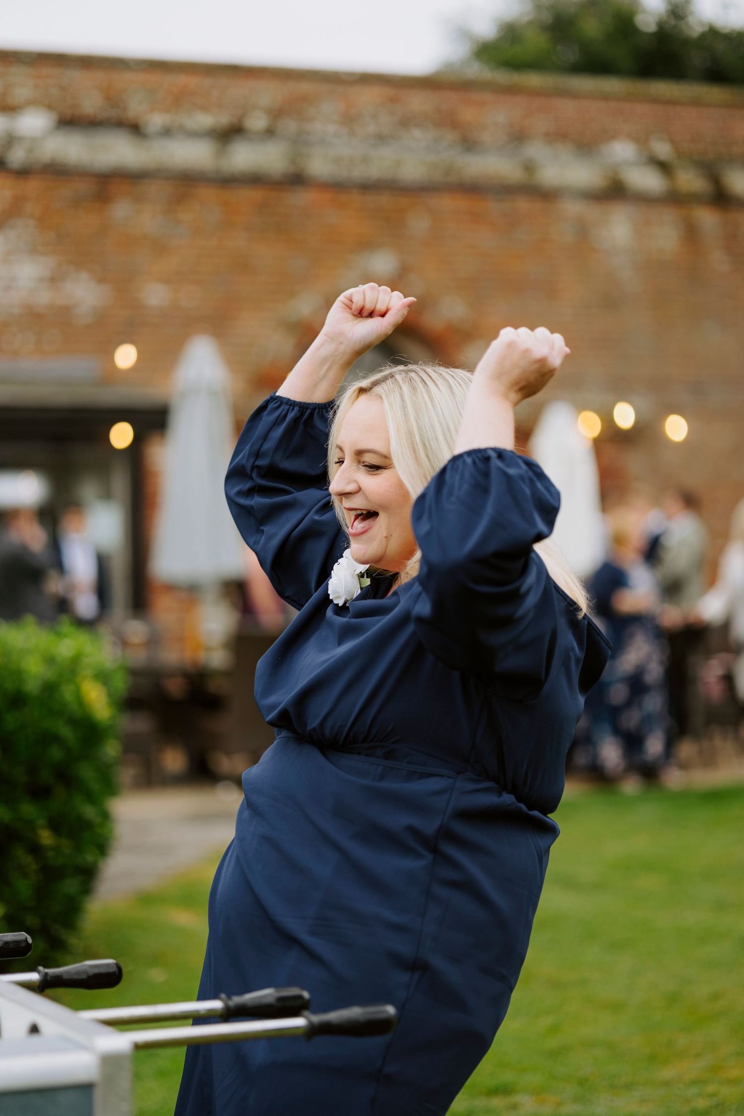 A woman in a navy dress jumps joyfully at an outdoor reception with brick walls and string lights.