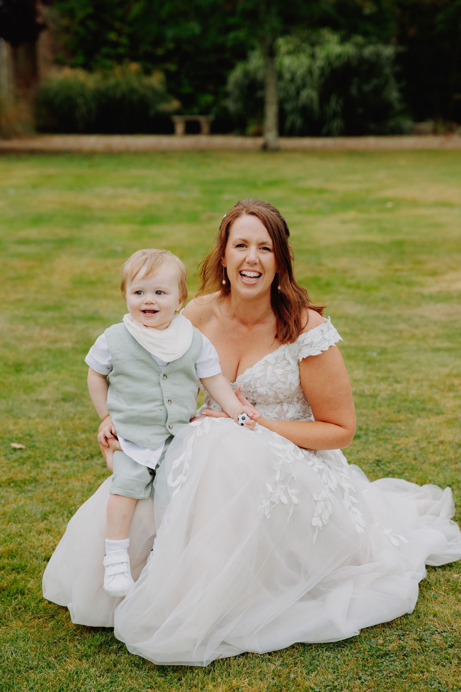 A bride in a white lace dress sits on grass with a smiling toddler wearing a light colored outfit.
