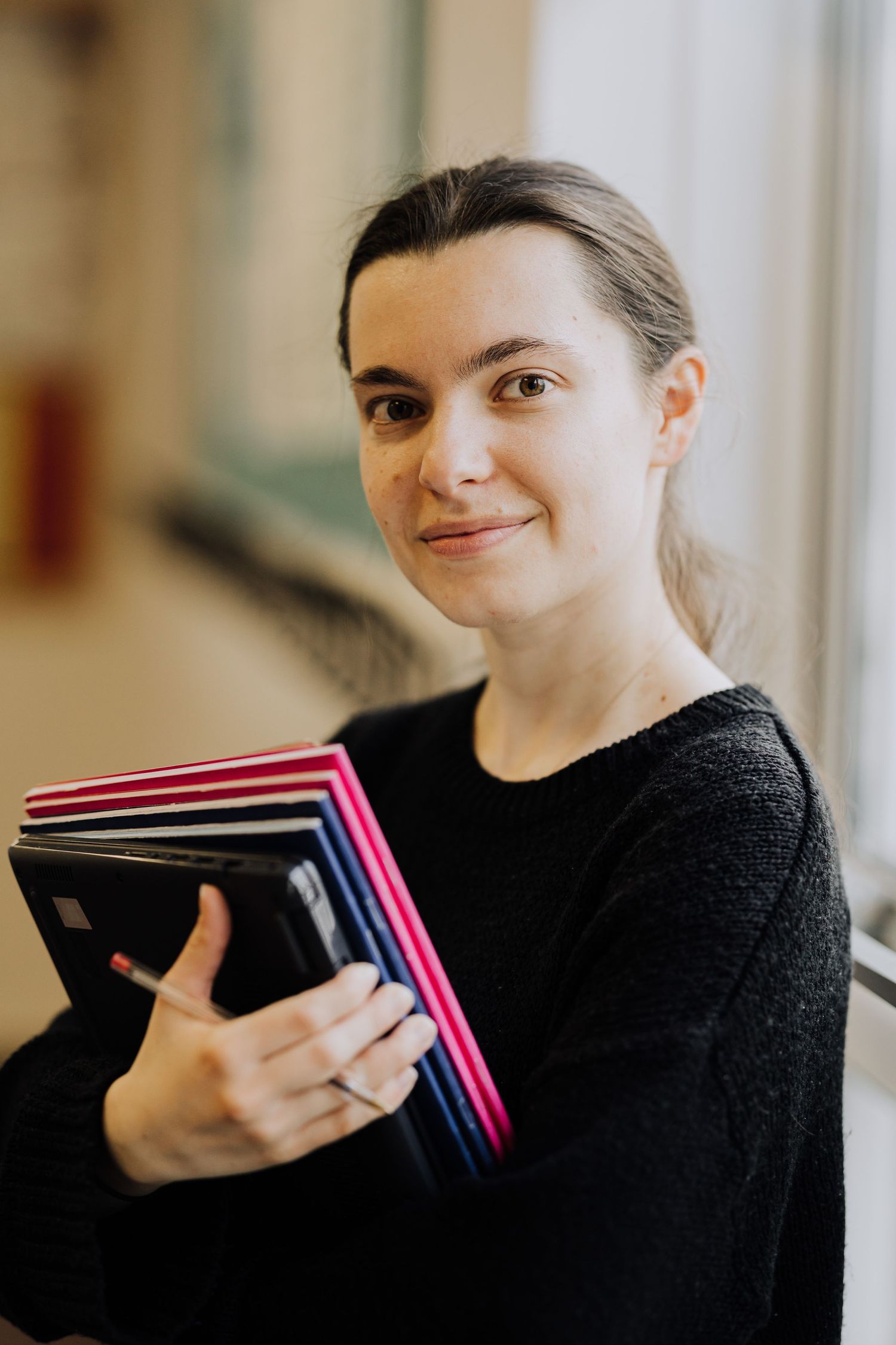 Someone in a black sweater smiling while looking at stacked notebooks with pink edges.