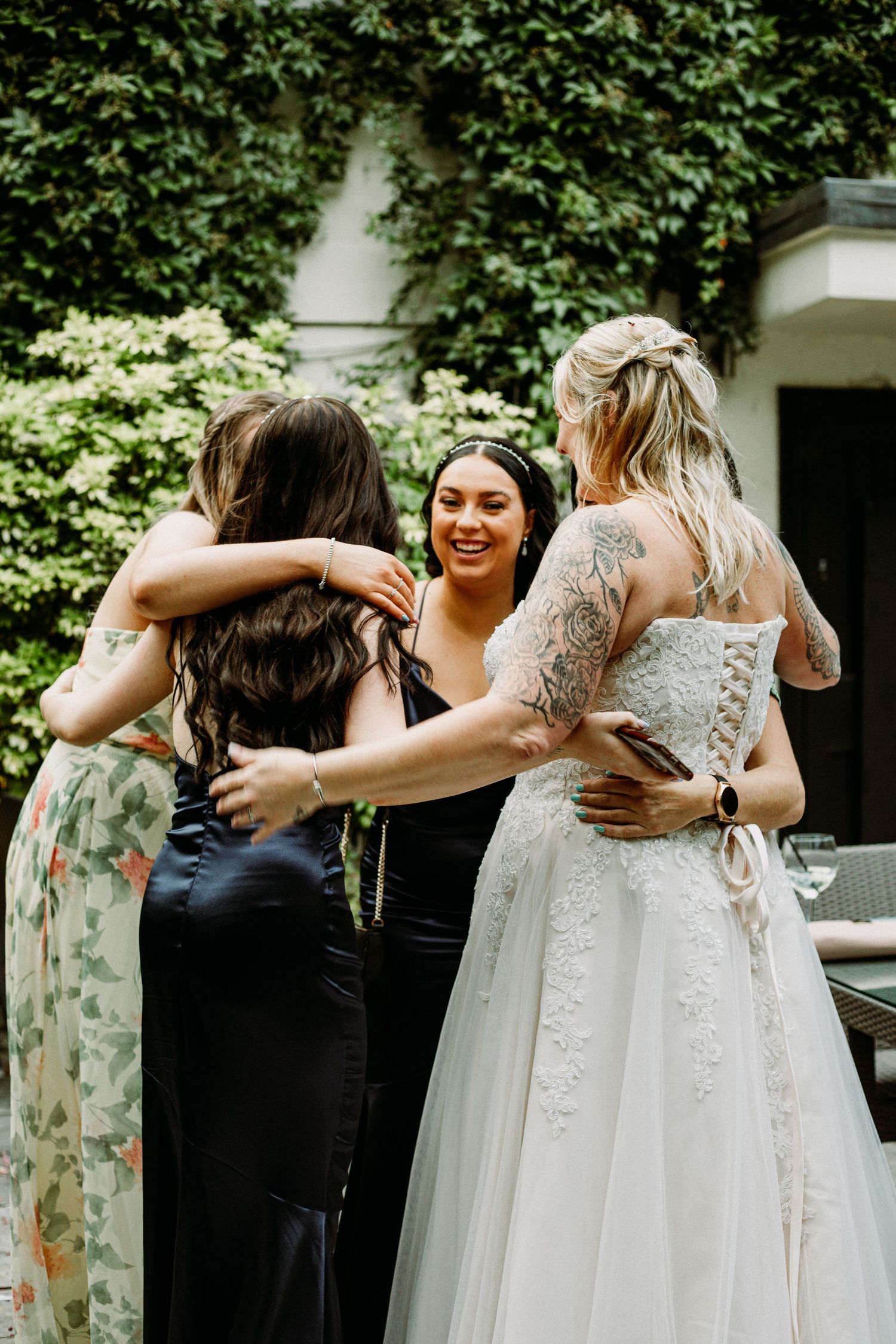 A group shares an emotional embrace at a wedding, with a bride in white lace and bridesmaids in floral and black dresses.