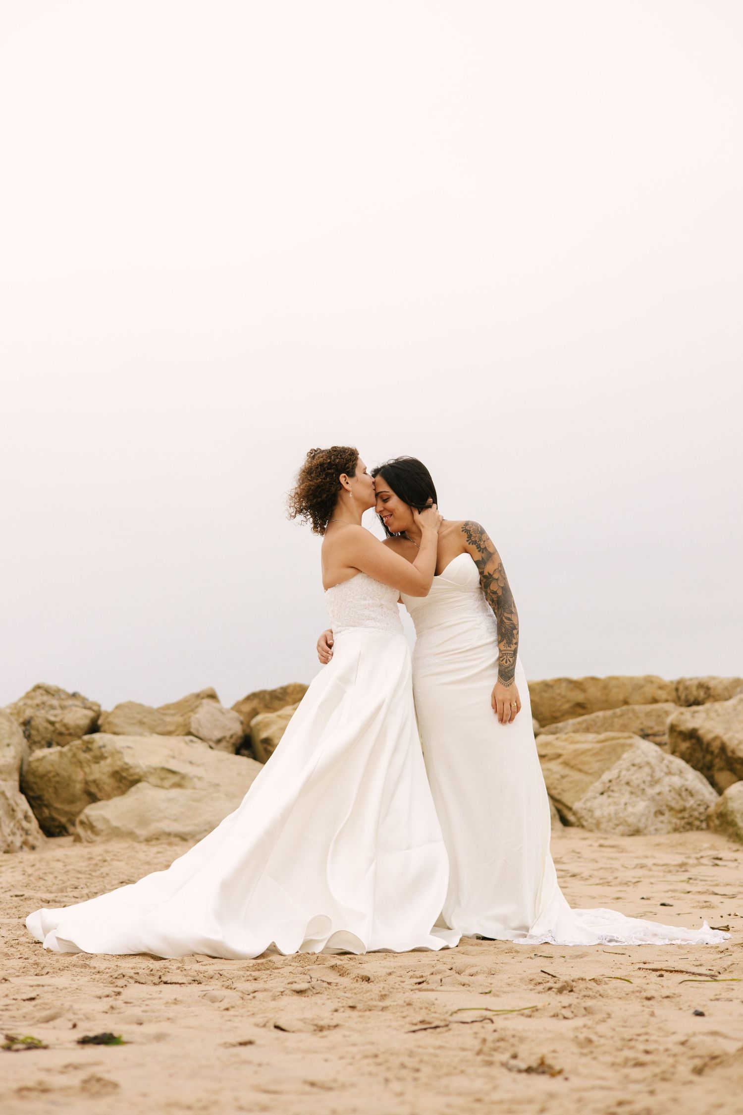 Two brides in white wedding dresses embrace on a rocky beach at sunset.