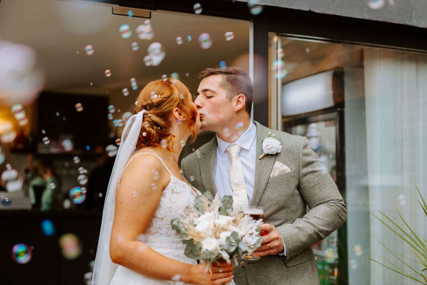 Couple shares romantic kiss surrounded by floating bubbles outside venue.