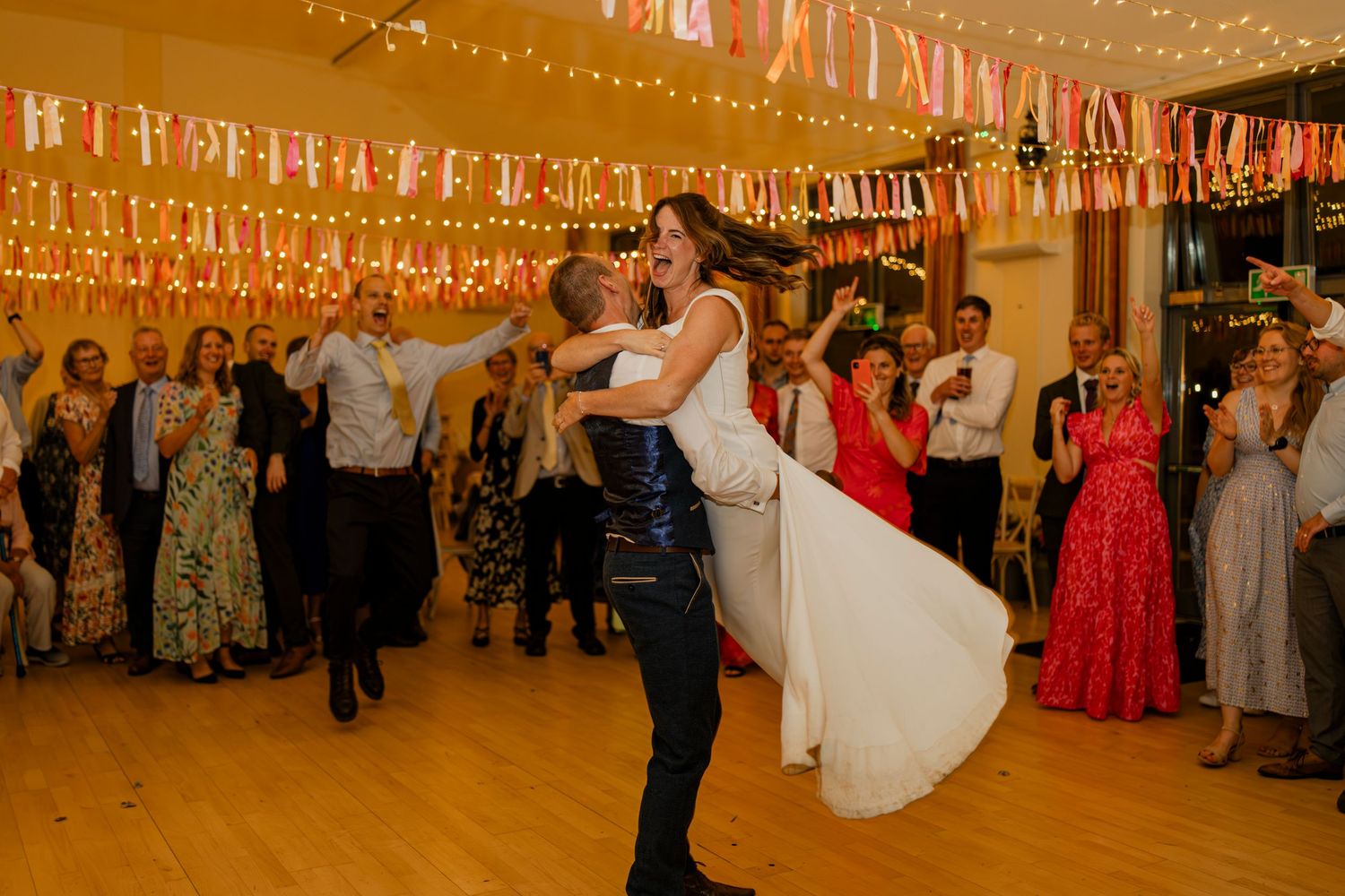 Wedding party celebrates under romantic string lights at indoor reception with guests dancing and socializing.