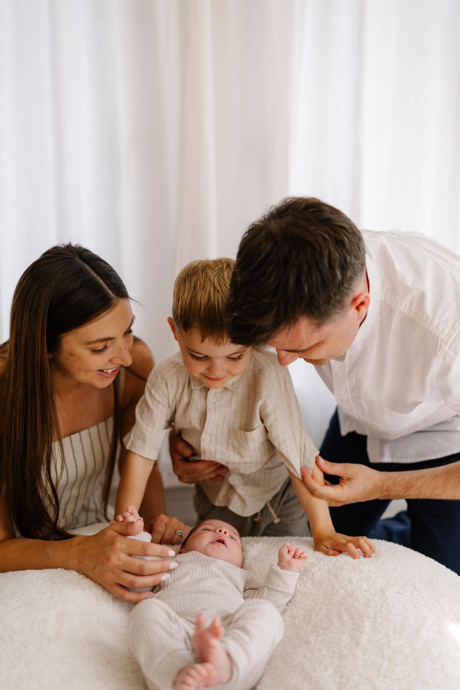 Parents in white shirts leaning over white bedding while gazing lovingly at their baby.