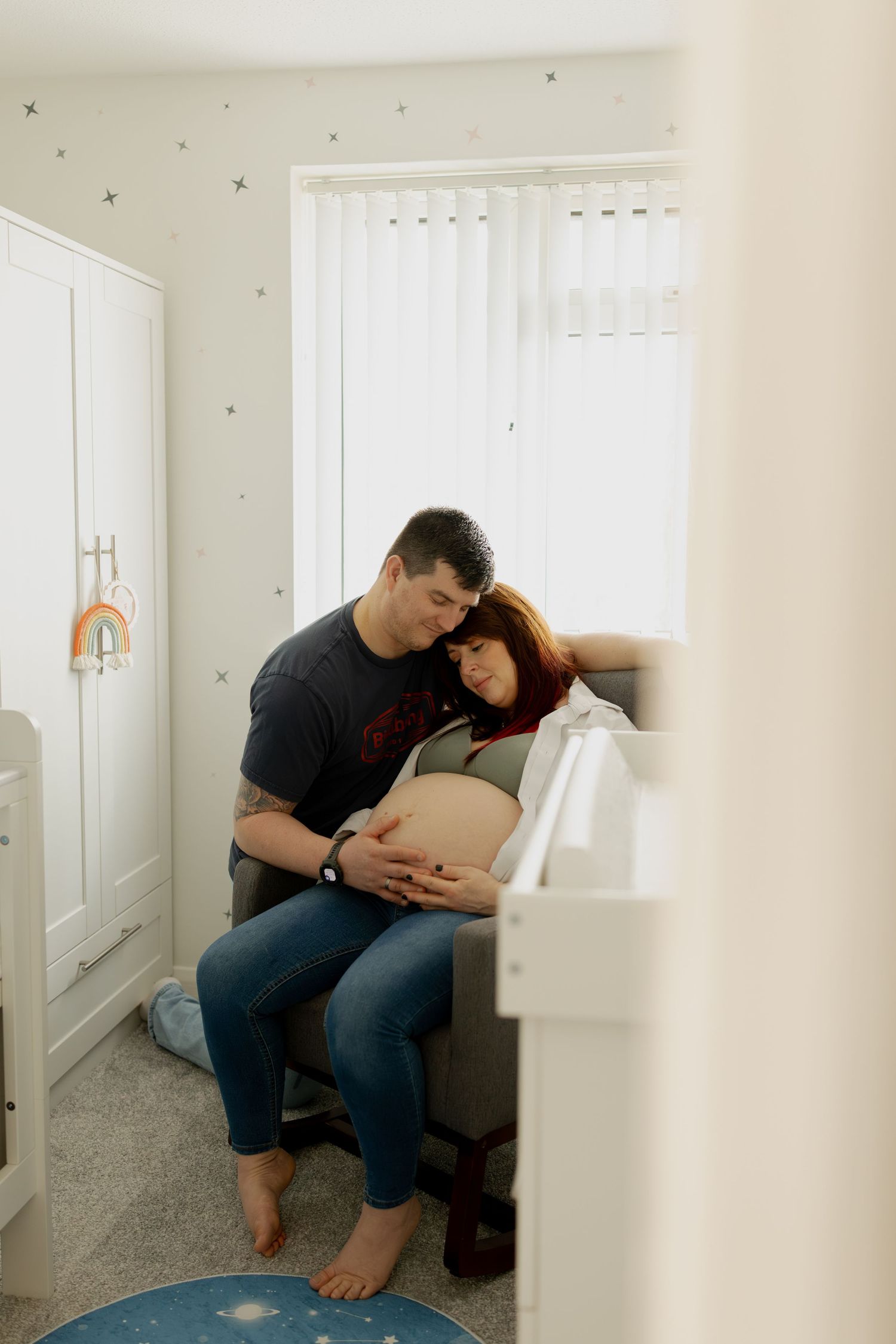 A parent sits with a baby in a cozy corner of a minimalist white nursery room.