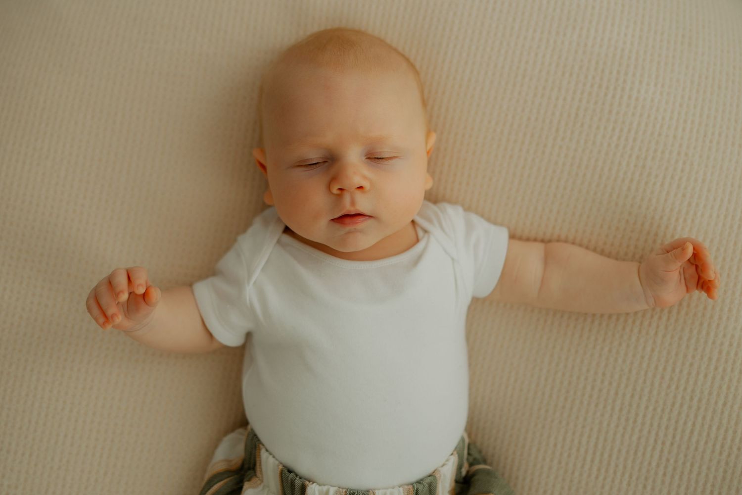 Baby lying on a beige textured surface wearing a white onesie with arms outstretched.
