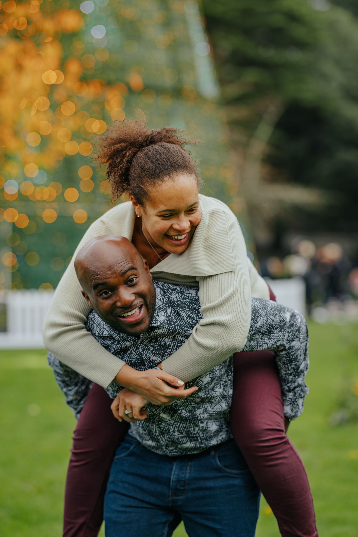 A playful couple laughs together during a piggyback ride in a park with twinkling lights in the background.