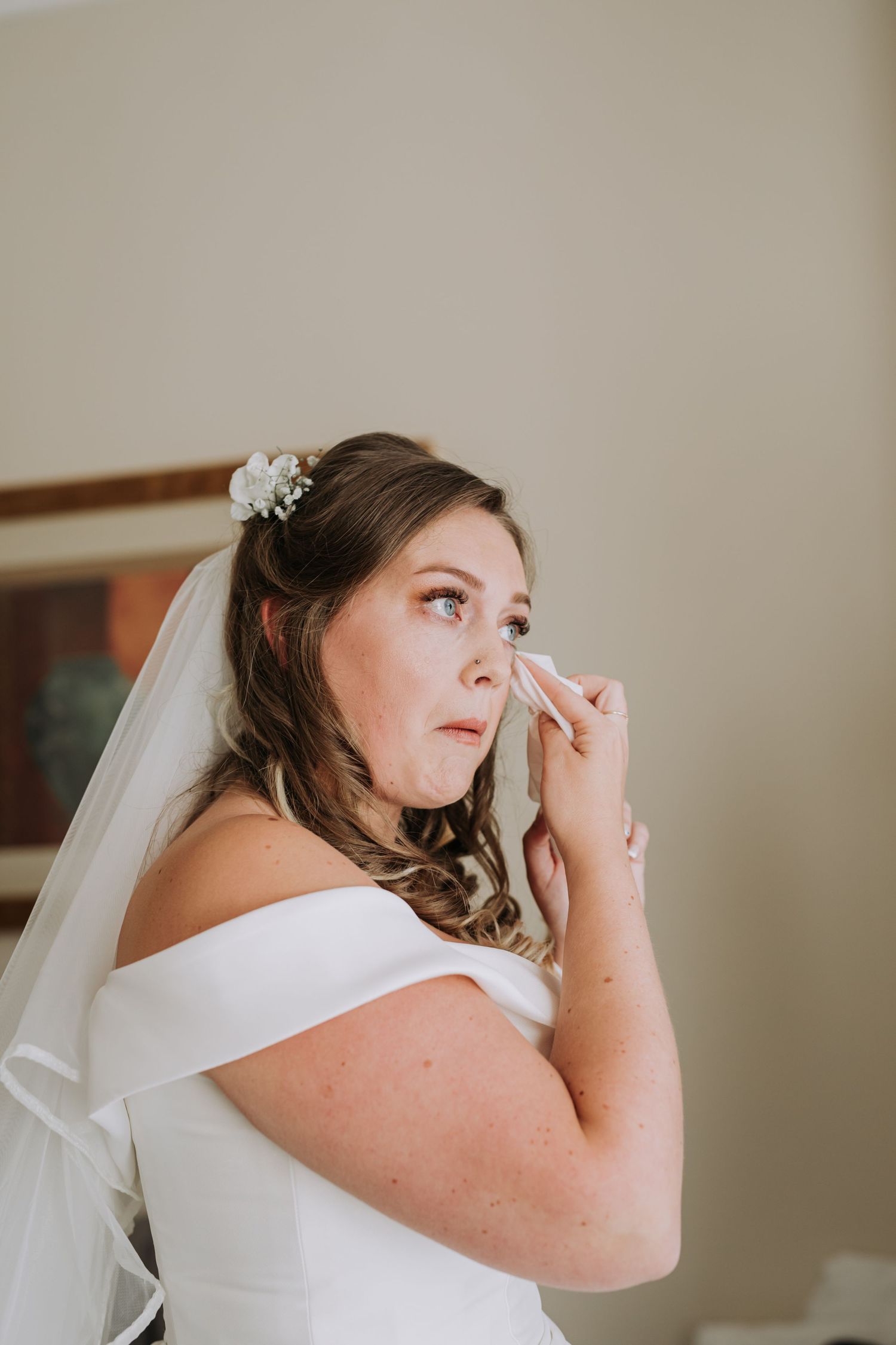Emotional bride in white dress and veil wipes tears while getting ready.