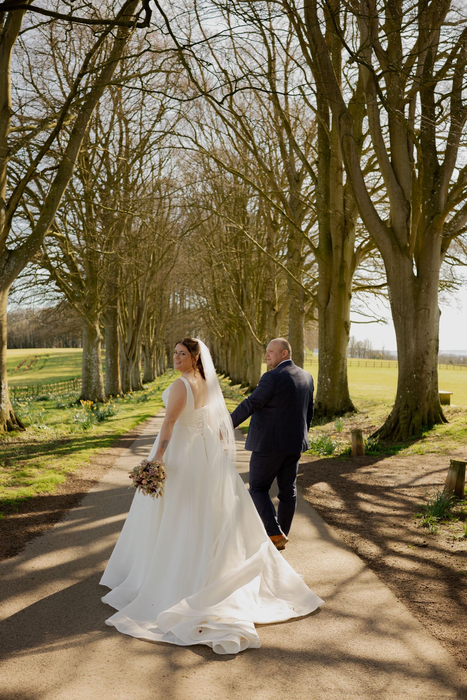 Bride and groom walking along a tree-lined path, bride in flowing white gown with veil, groom in navy suit.