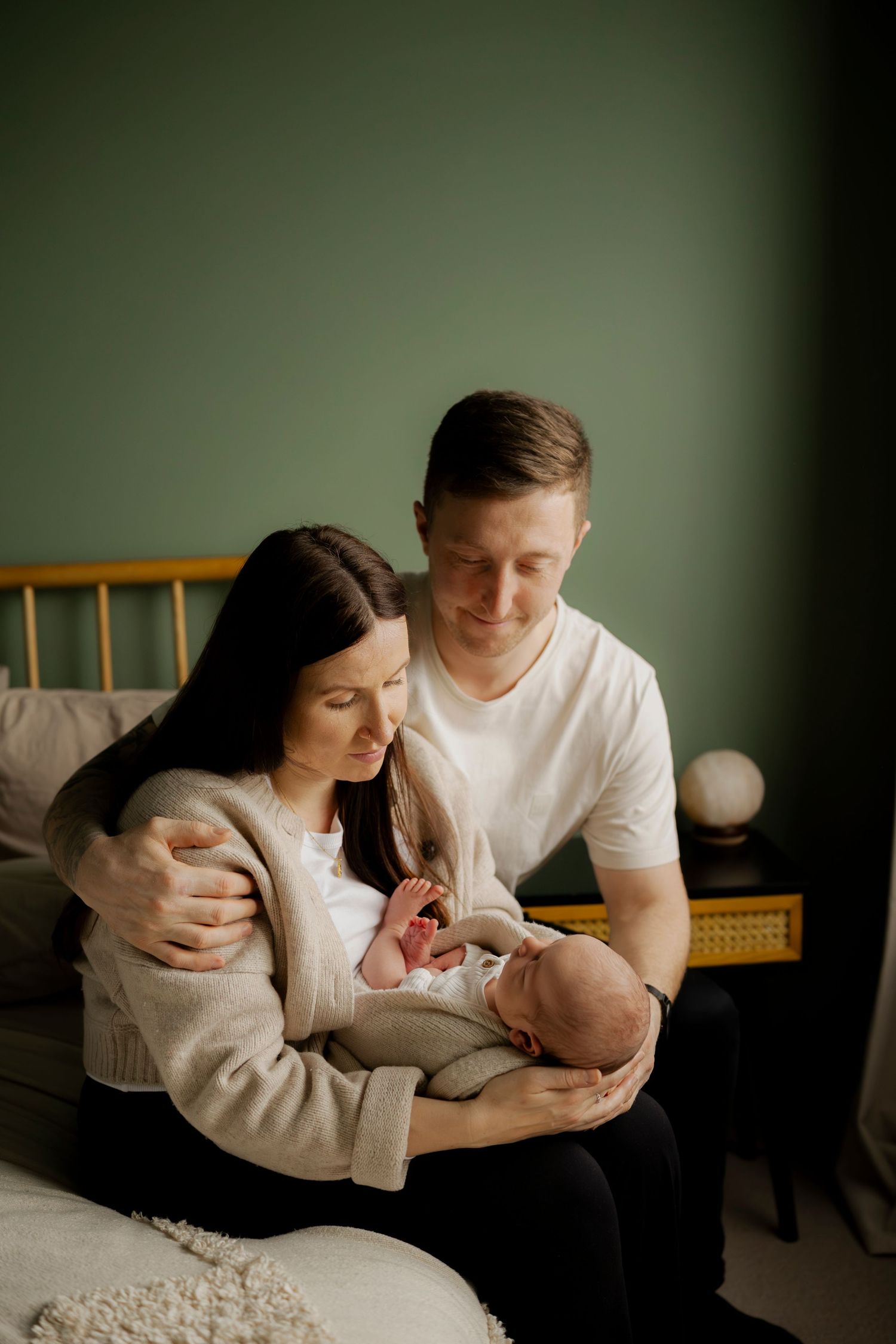 A couple in white and black clothing share tender moments with their newborn baby in a softly lit room.