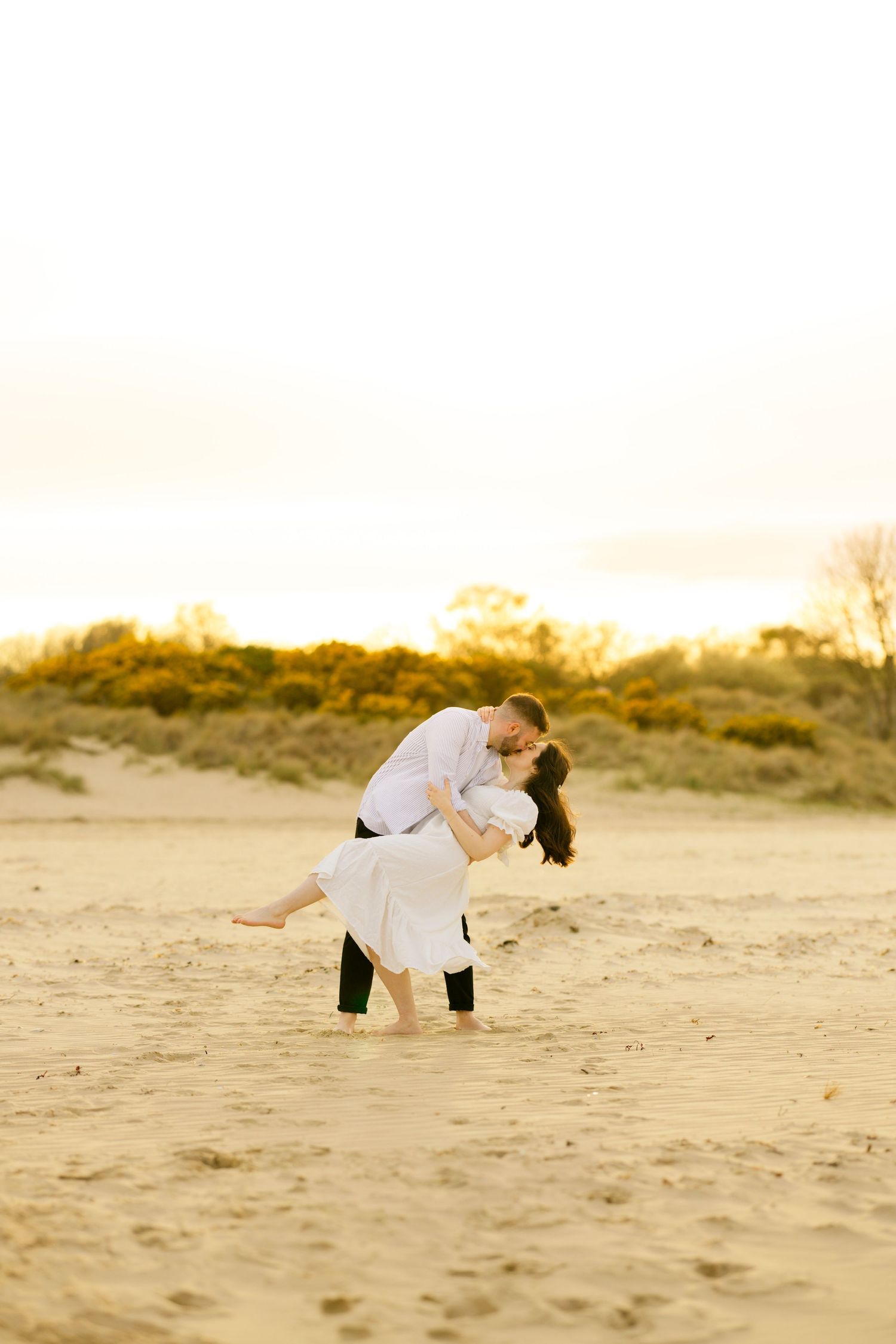 Dancing couple in white attire sharing intimate moment on sandy beach at sunset.