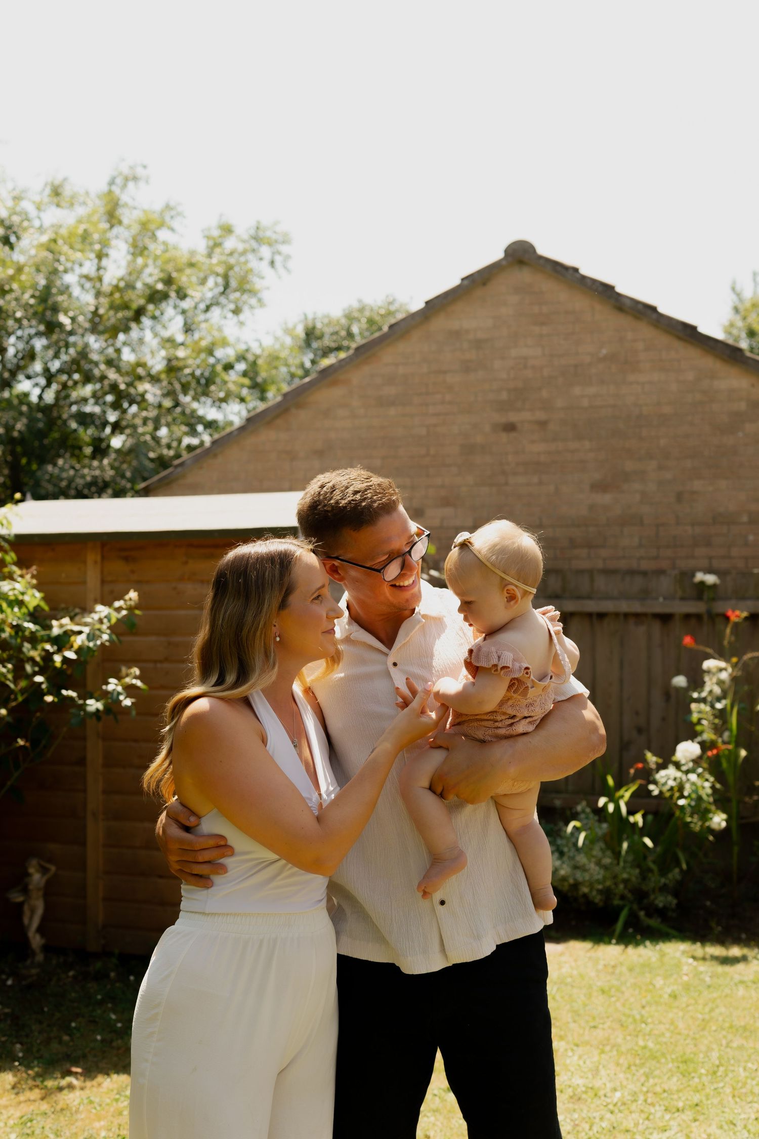A young couple smiling at their baby girl in a sunny backyard garden setting.