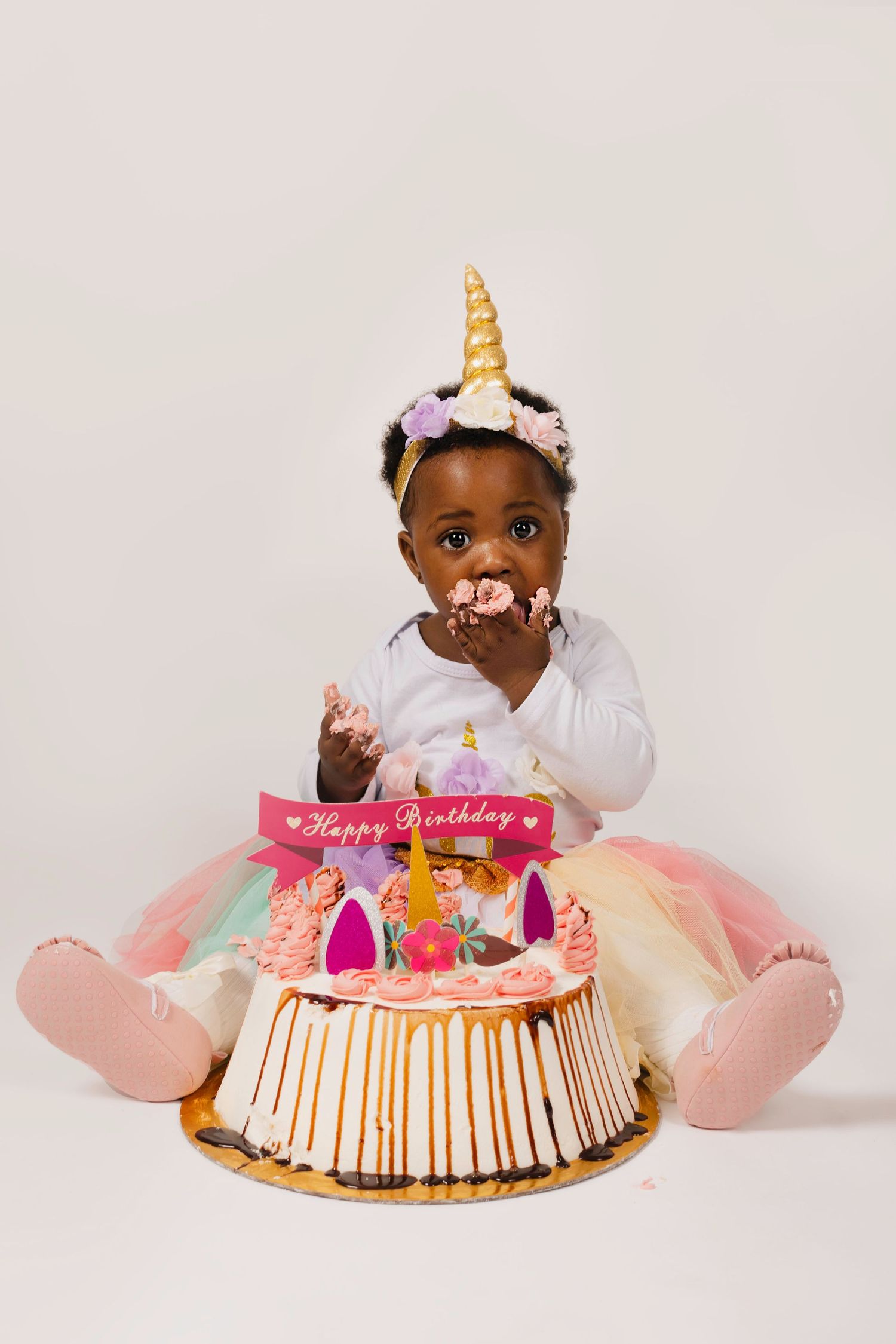 Baby in unicorn headband and tutu dress celebrating first birthday with pink decorated cake.