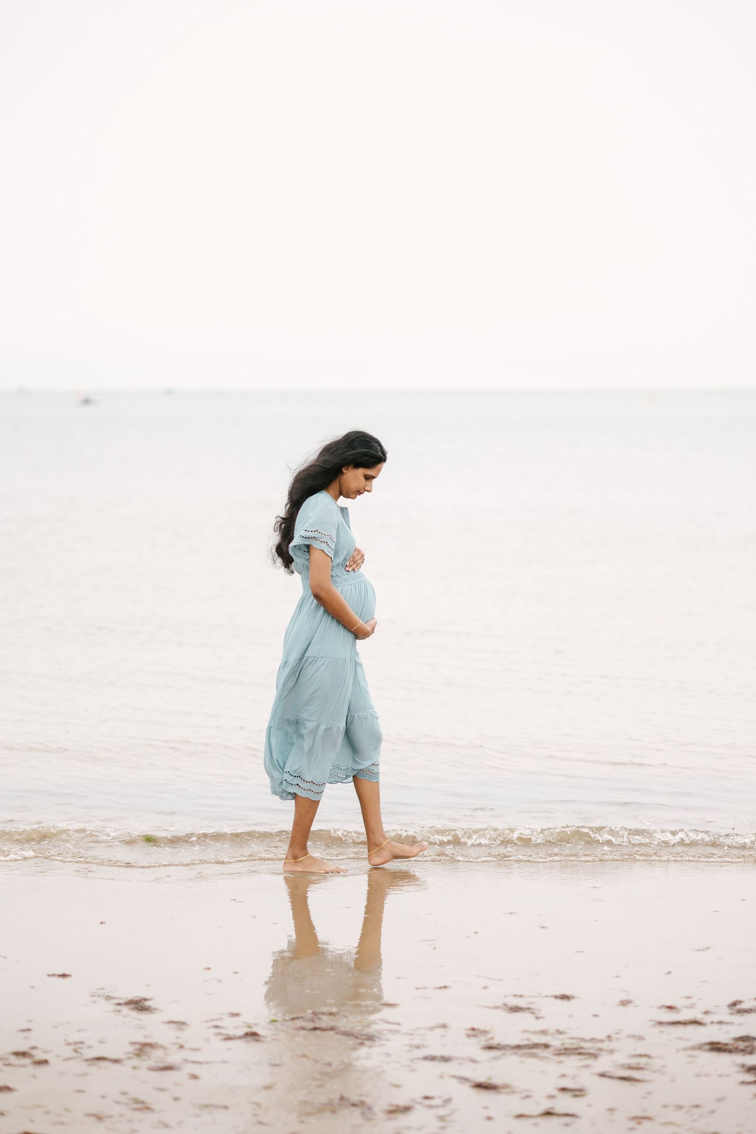 Pregnant woman in flowing blue dress walking barefoot on beach.
