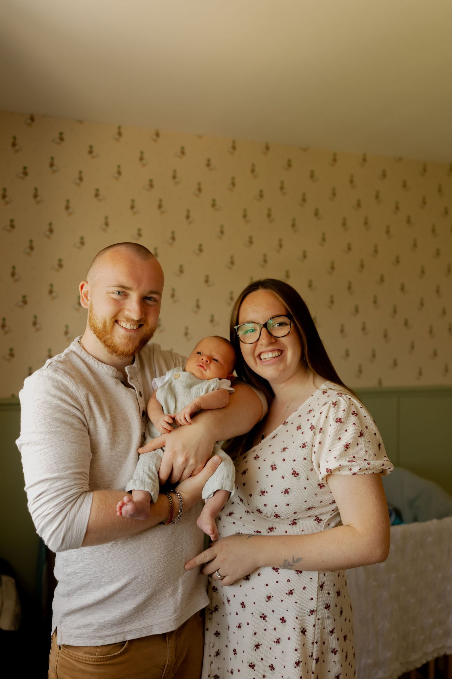 A smiling couple holds their newborn baby together in a warmly lit room with patterned wallpaper.