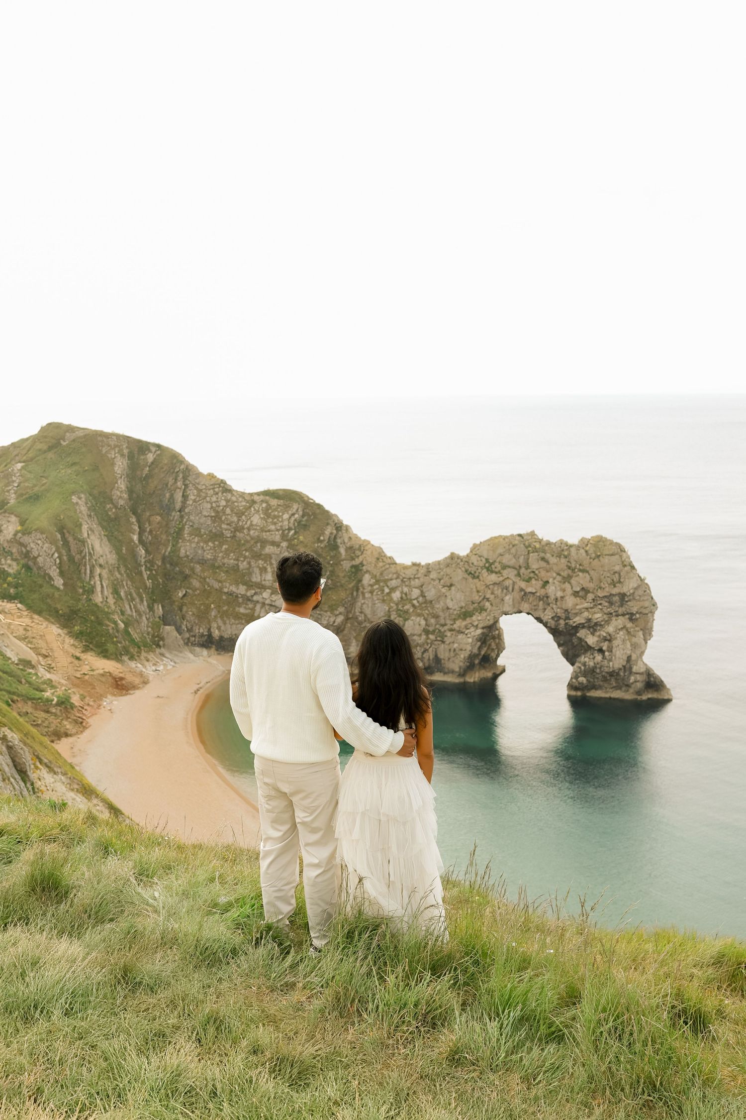 Couple in white outfits standing on cliffside overlooking Durdle Door arch and turquoise sea, Dorset, England.
