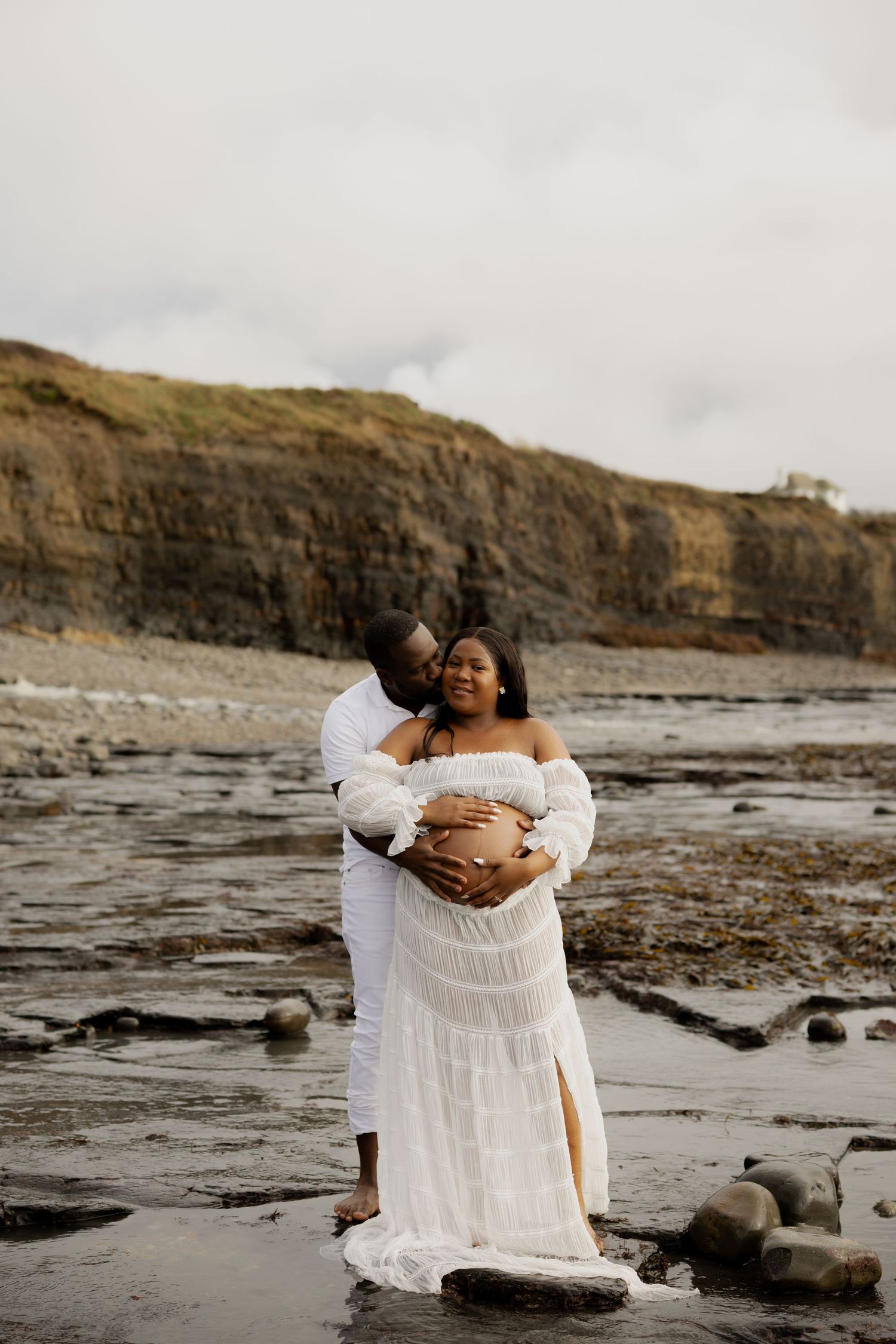 Couple in white outfits sharing a tender embrace on a rocky beach with coastal cliffs in the background.