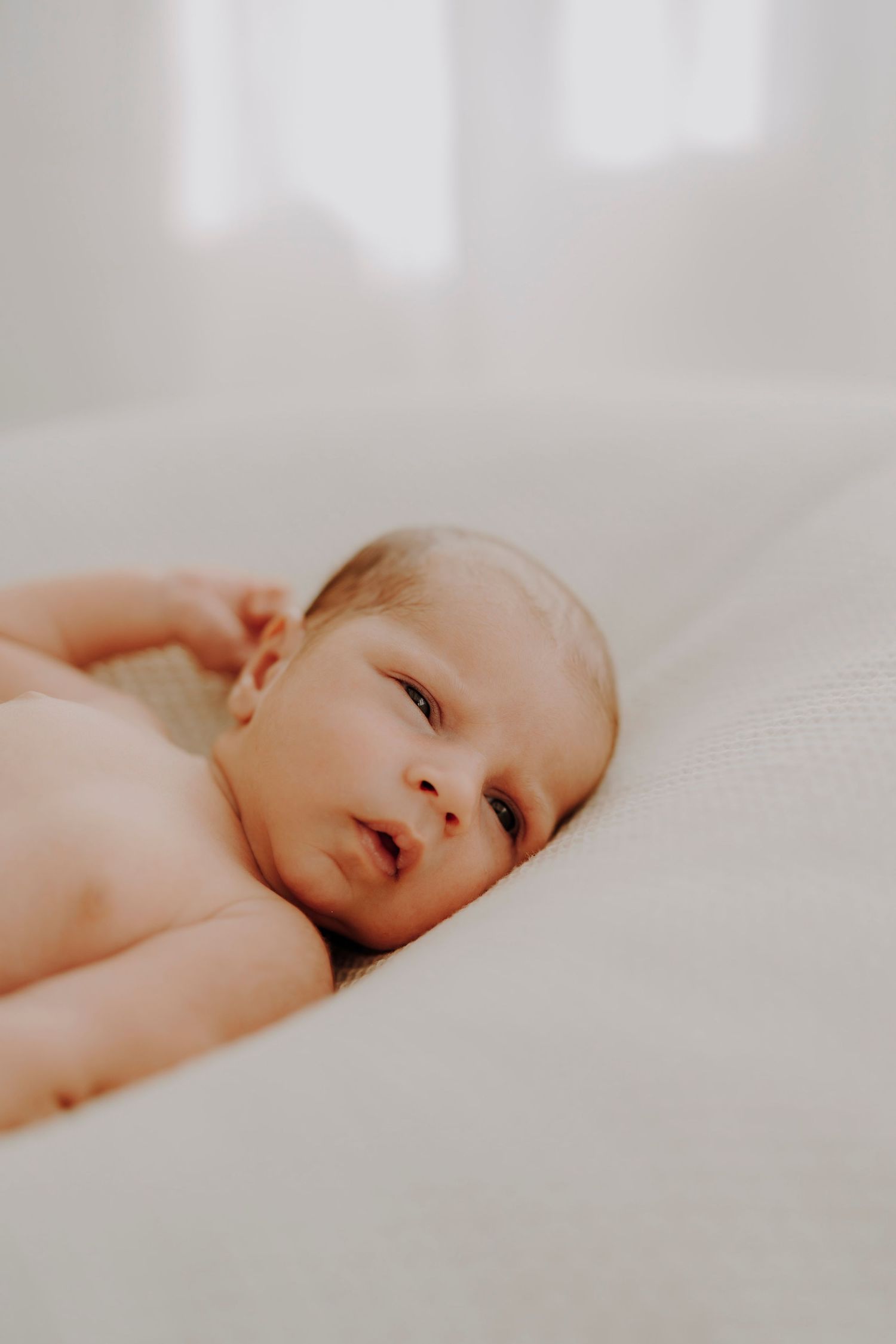 Newborn baby with peaceful expression lying on soft white bedding in natural light.
