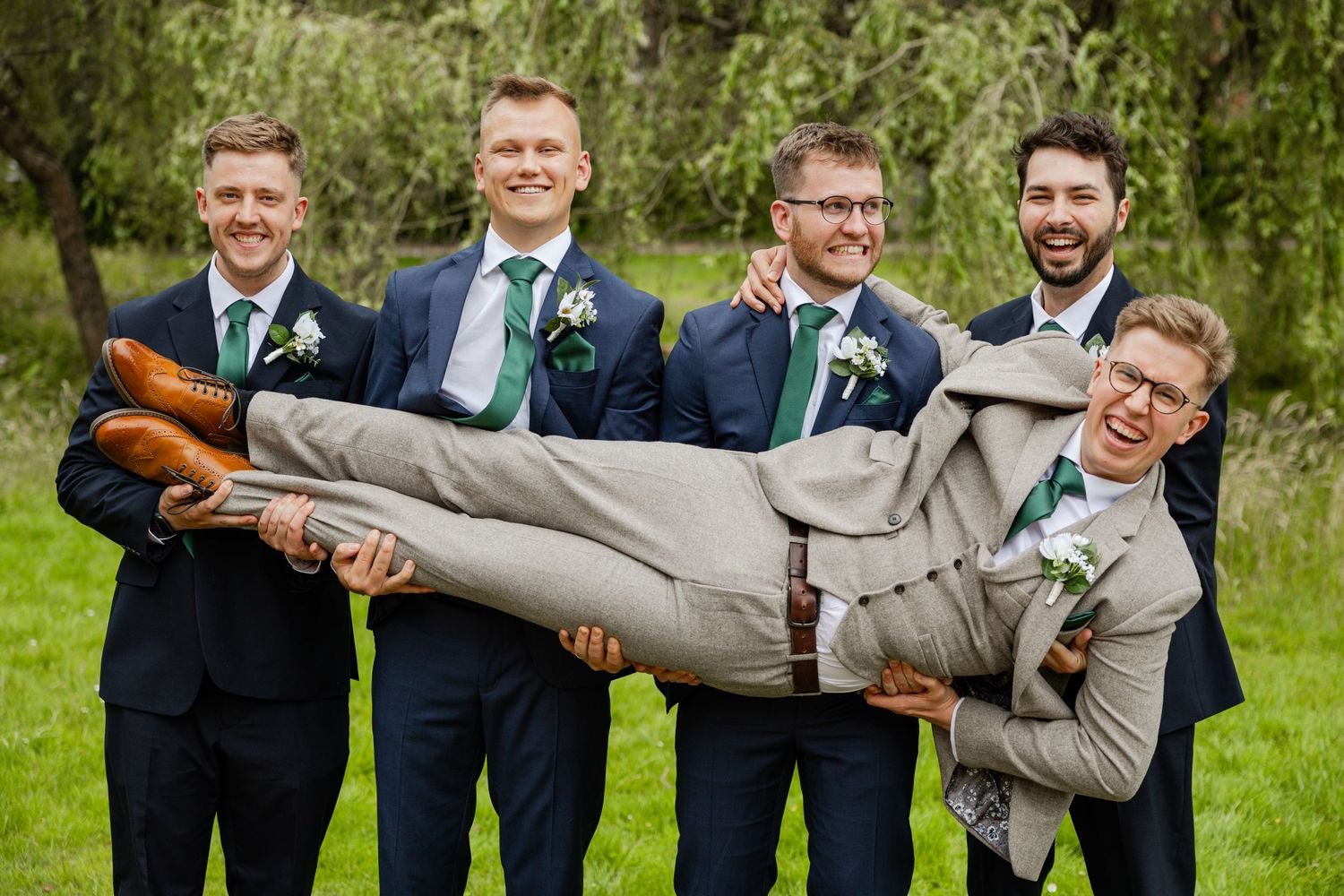 Groomsmen in matching suits playfully lifting and carrying a person horizontally outdoors.