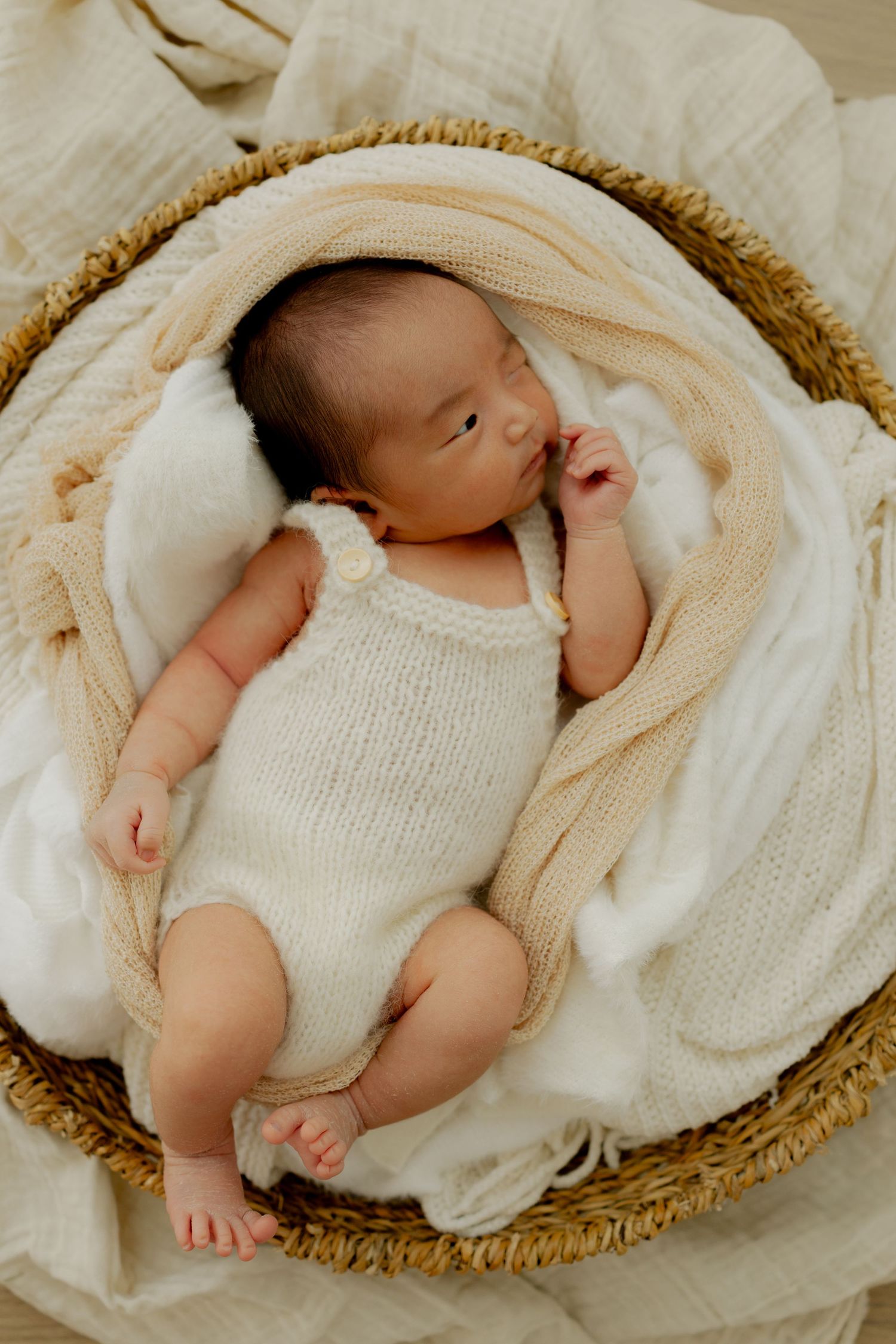 Newborn baby in cream knit romper sleeping peacefully in a woven basket with soft white and beige blankets.