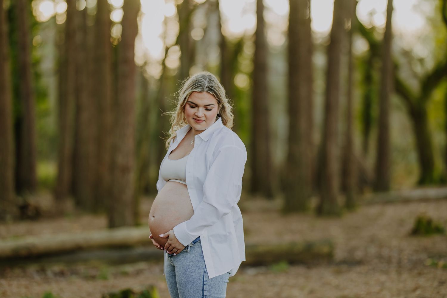 Pregnant woman in white top and jeans poses in a wooded area during golden hour.