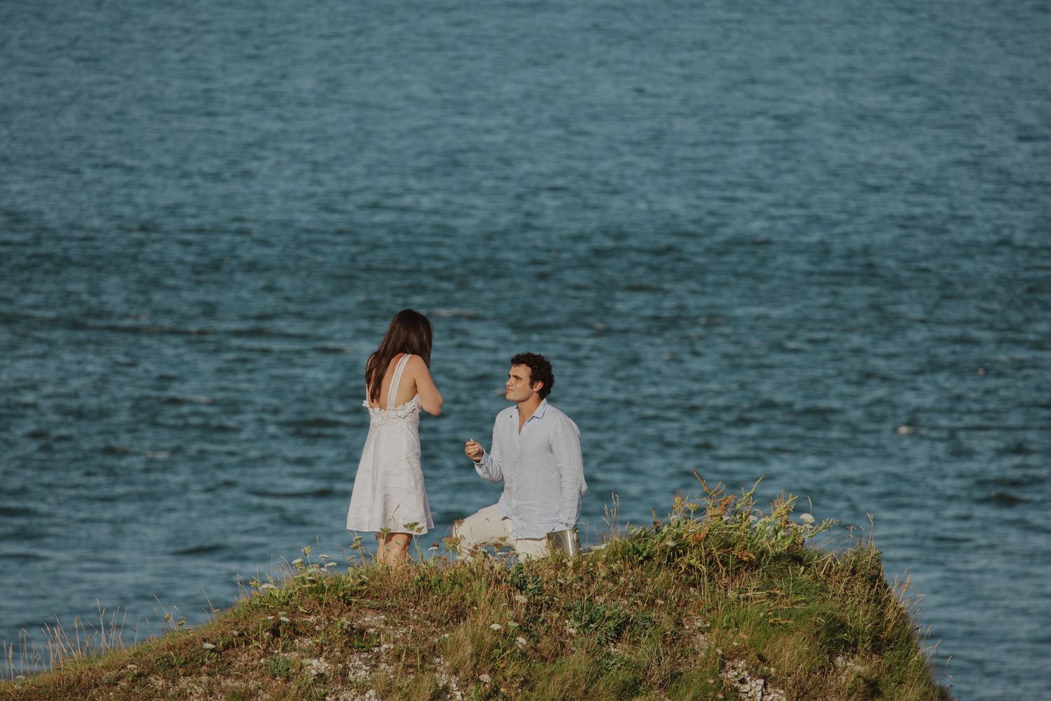 Marriage proposal scene on coastal cliff overlooking ocean at sunset.