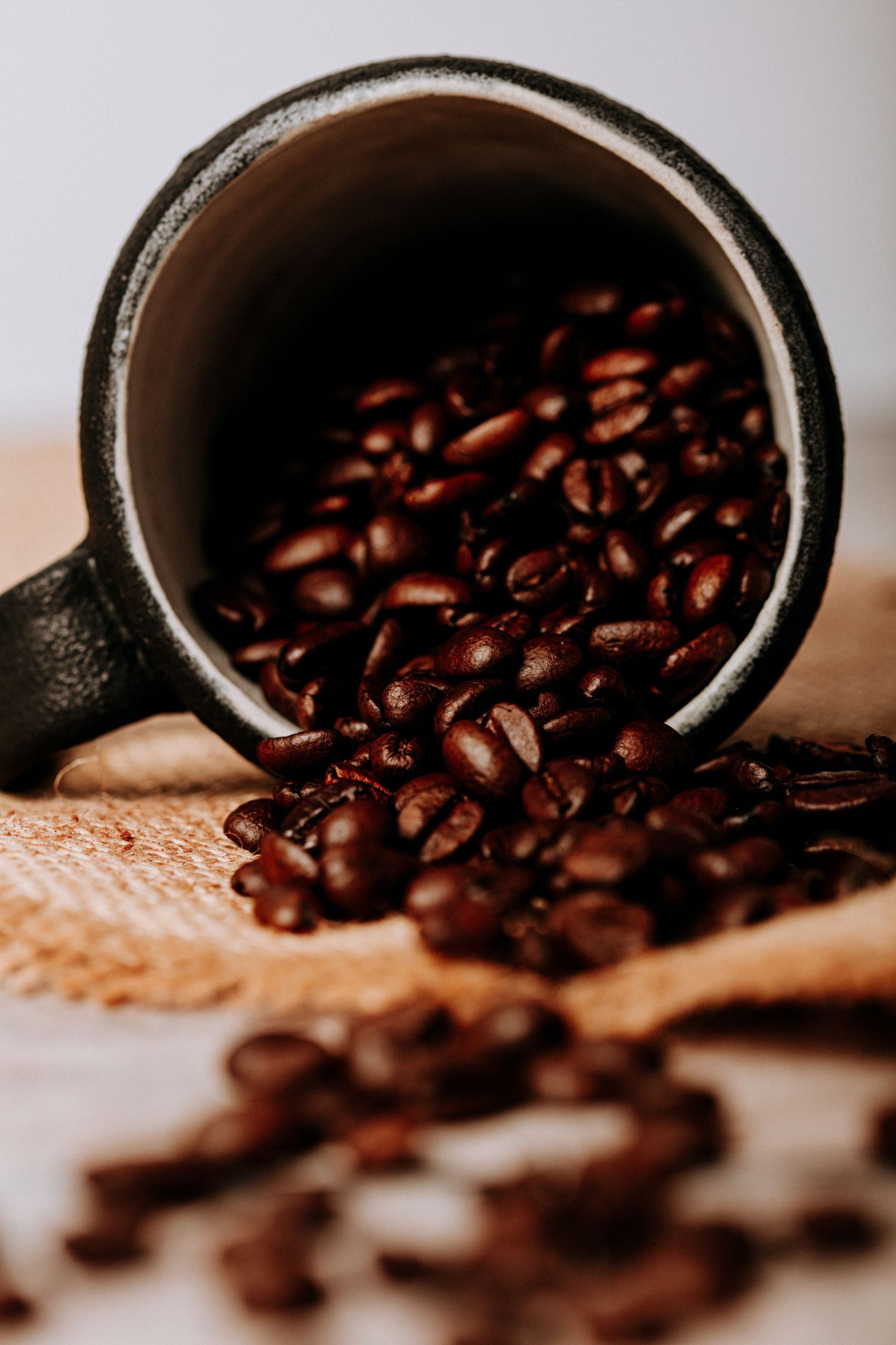 Close-up of fresh roasted coffee beans spilling from a dark ceramic cup onto burlap surface in moody lighting.