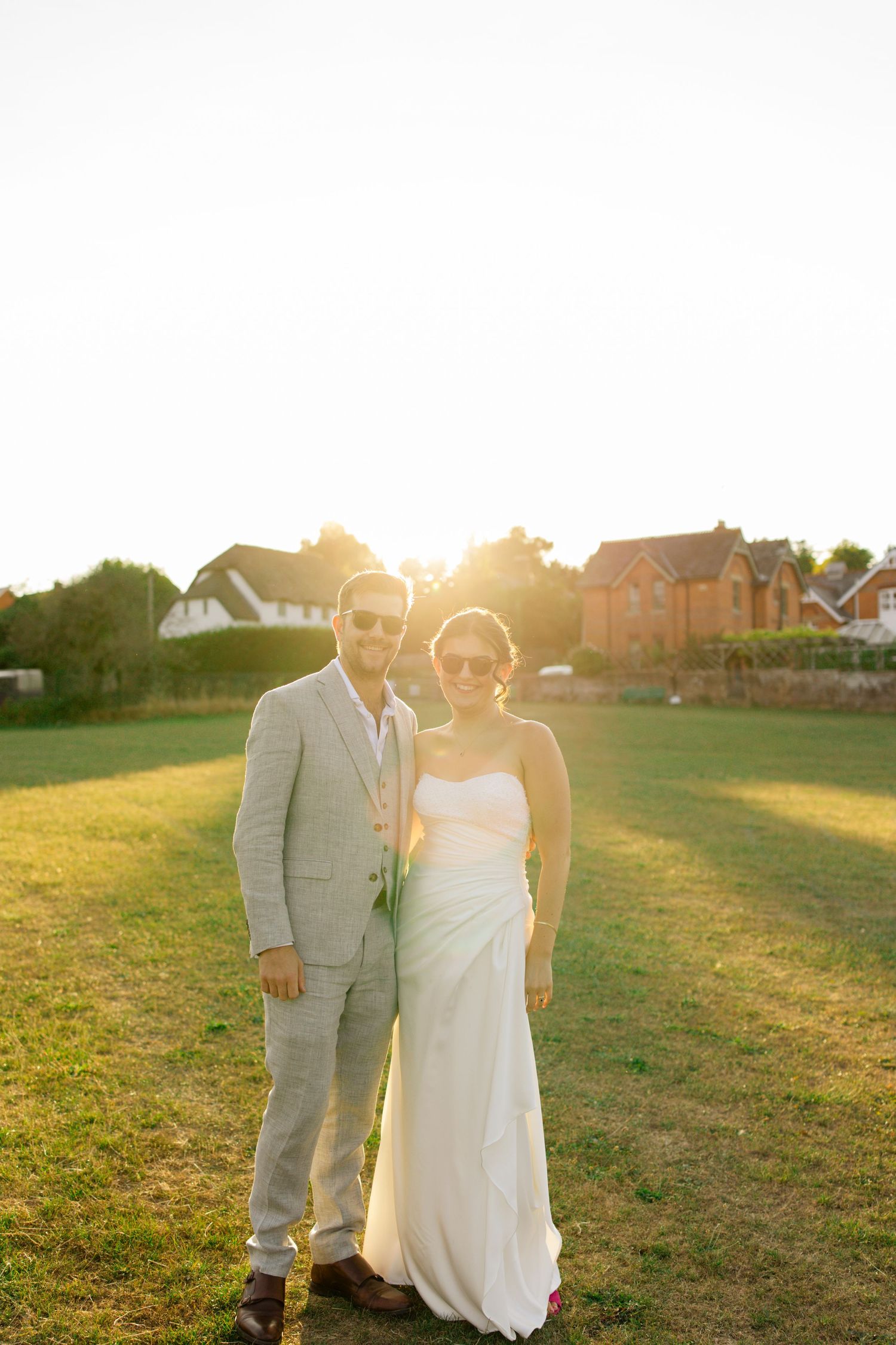 A couple walks hand in hand through a sunlit meadow during golden hour with homes in the background.