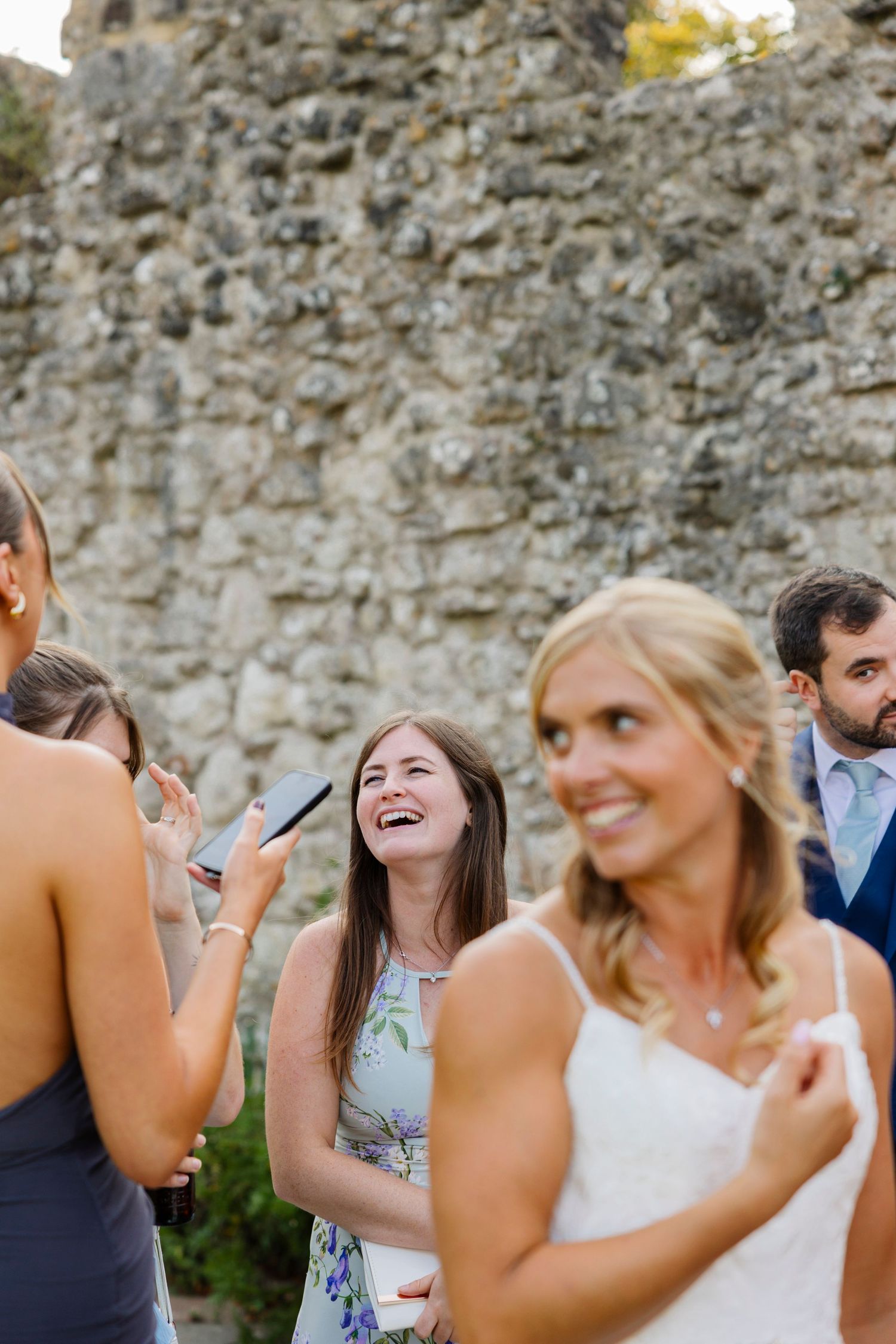 Wedding guests share joyful laughter during outdoor celebrations near stone walls.
