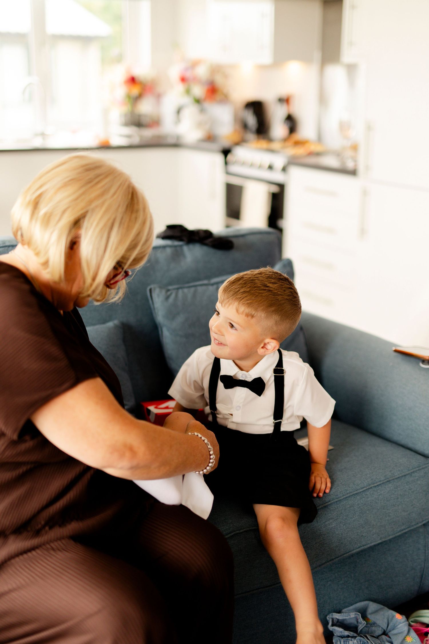 A young boy in suspenders and bowtie sitting on couch during wedding preparations.