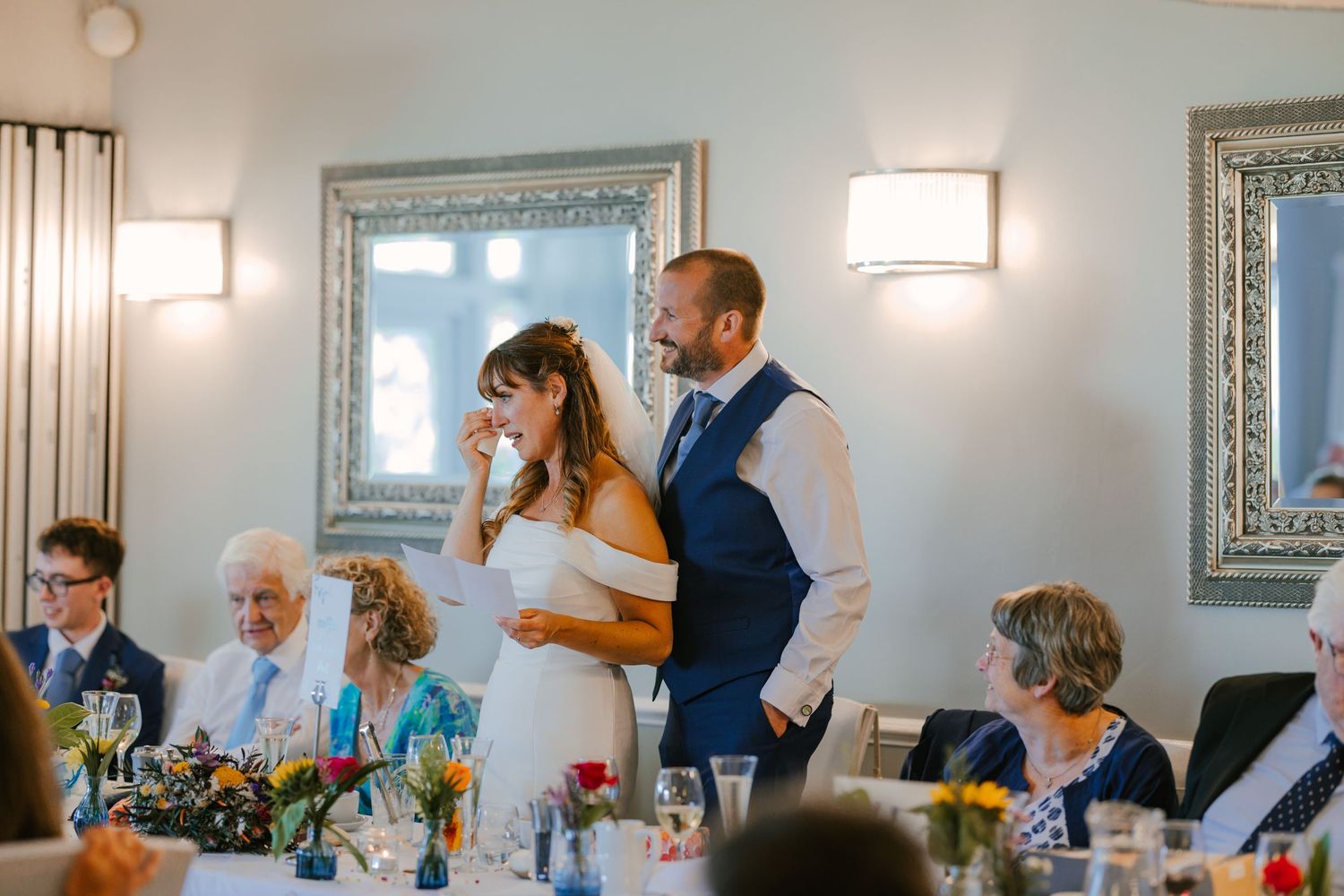 A couple delivers wedding speeches at their reception with elegant silver-framed mirrors in background.