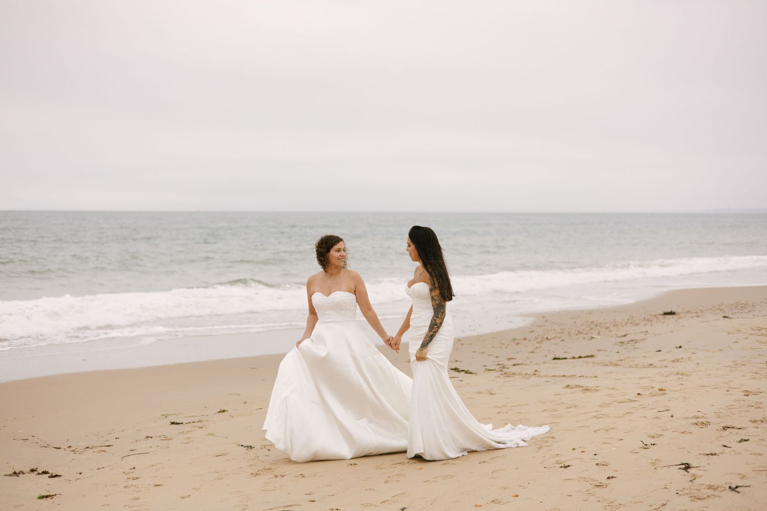 Two brides in white gowns hold hands while walking along a peaceful beach.
