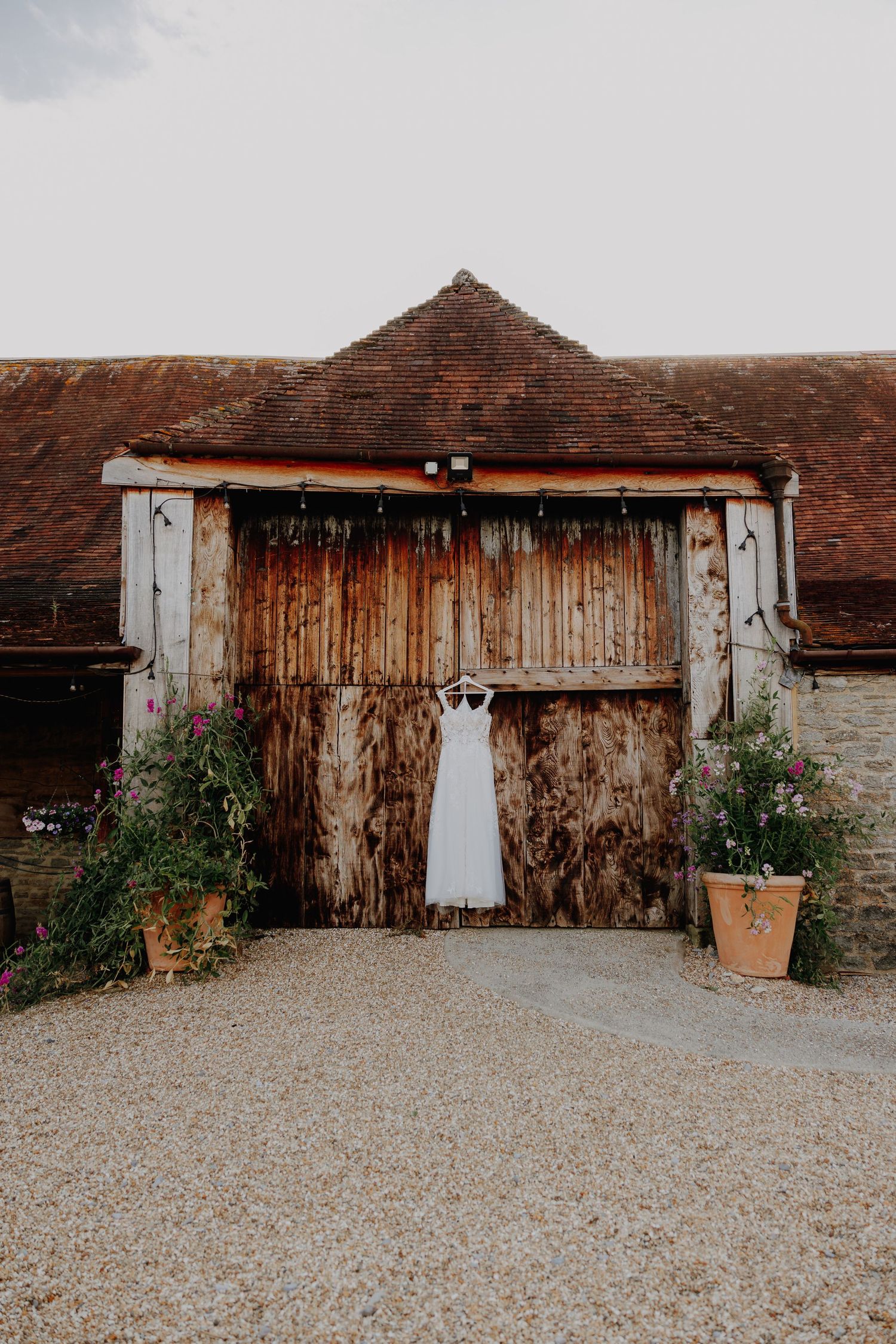 A white wedding dress hangs on rustic wooden barn doors with terracotta pots on either side.