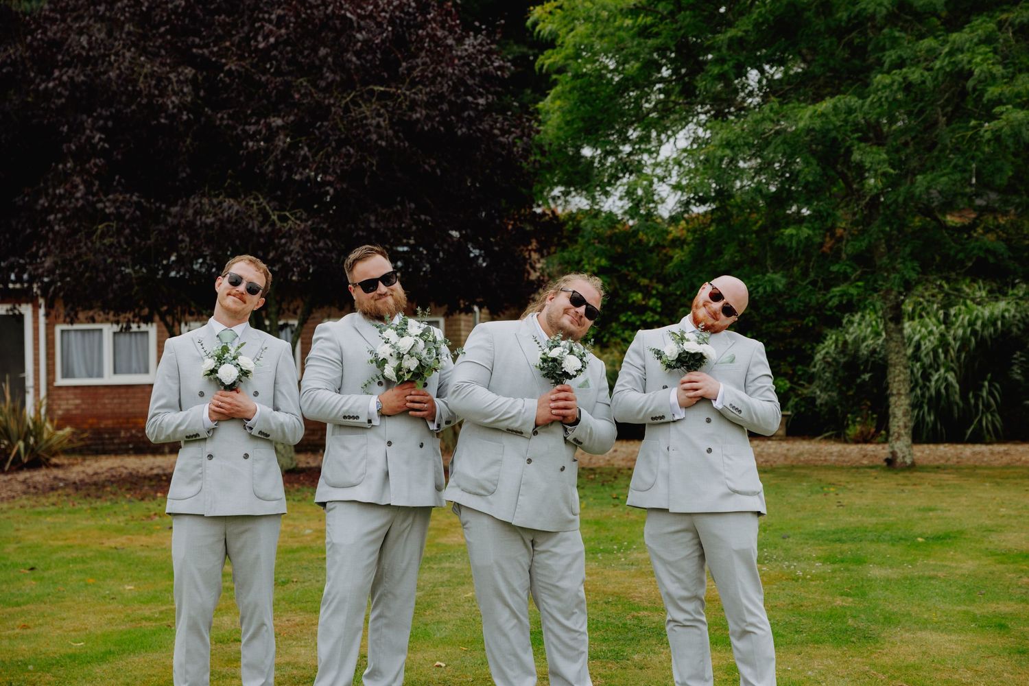 Four groomsmen in light gray suits pose with drinks while making playful expressions.
