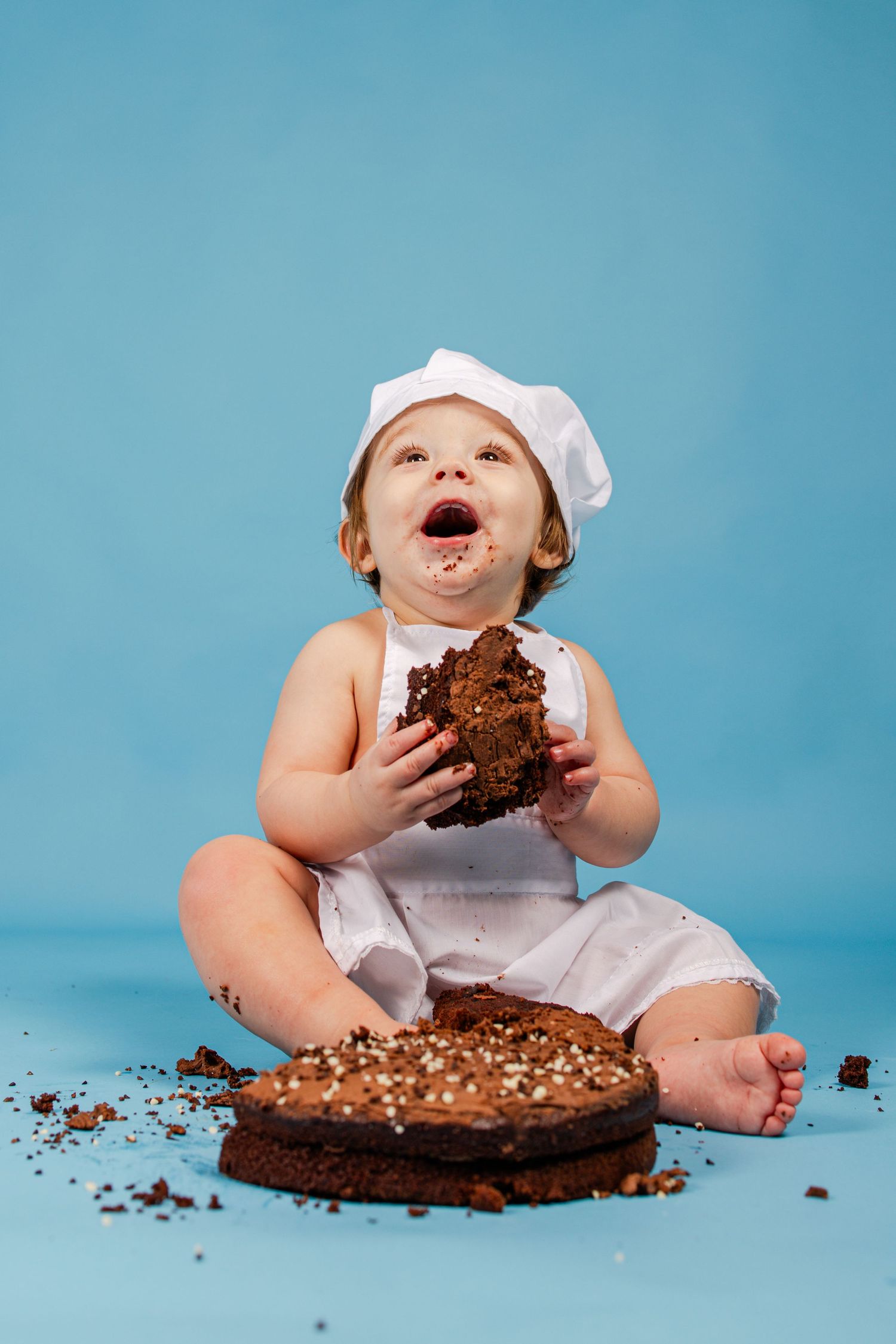 Messy baby in chef hat enjoying chocolate cake during cake smash photoshoot on blue background.