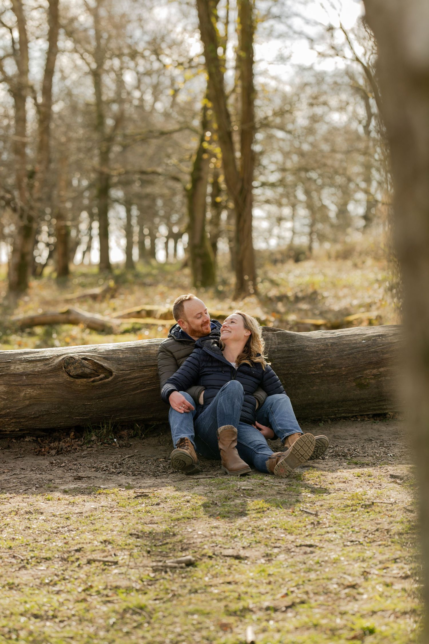 Couple sharing laughter while sitting on fallen log in sunlit forest.