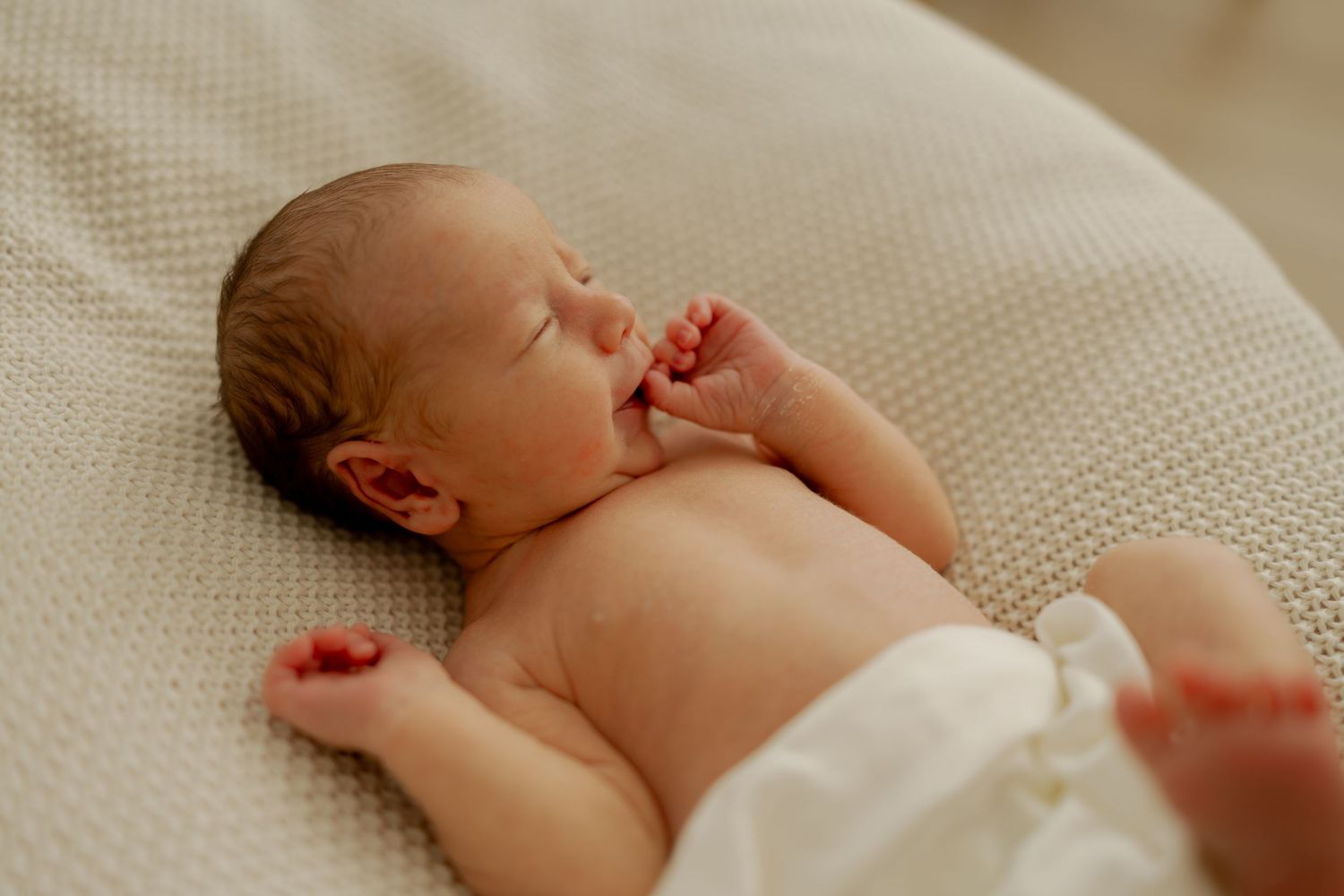 A sleeping newborn baby lies peacefully on soft white bedding while being gently touched by adult hands.