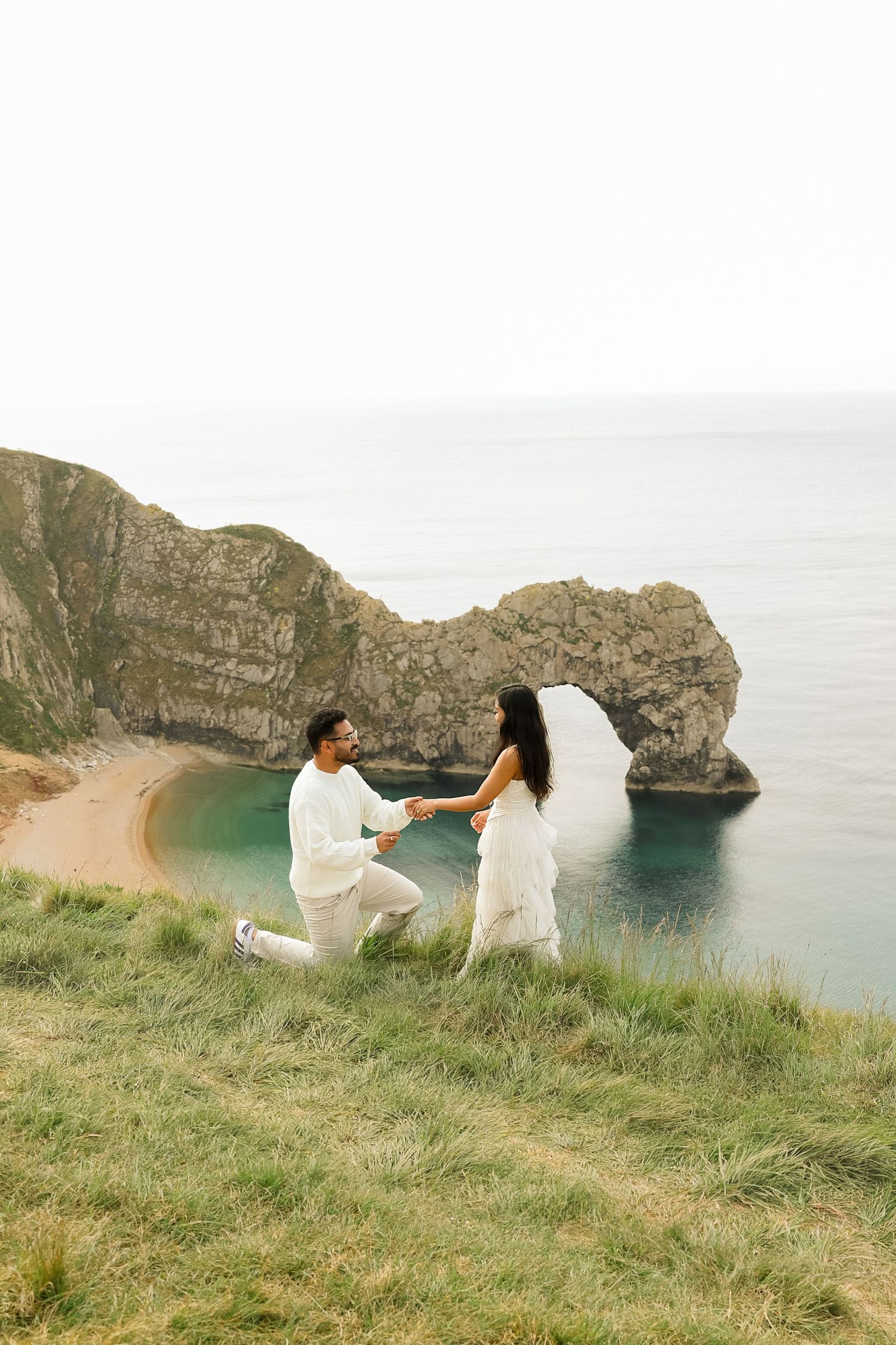 Couple in white attire at Durdle Door arch, Dorset, man on one knee proposing on grassy cliff overlooking turquoise sea.