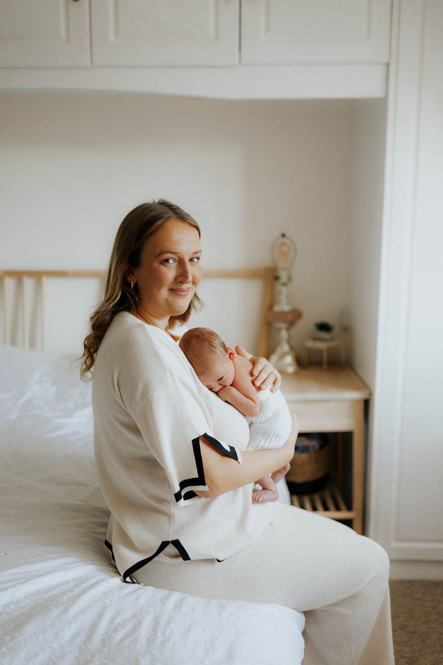 Mother in white sweater holding sleeping newborn while sitting on bed in bright bedroom.