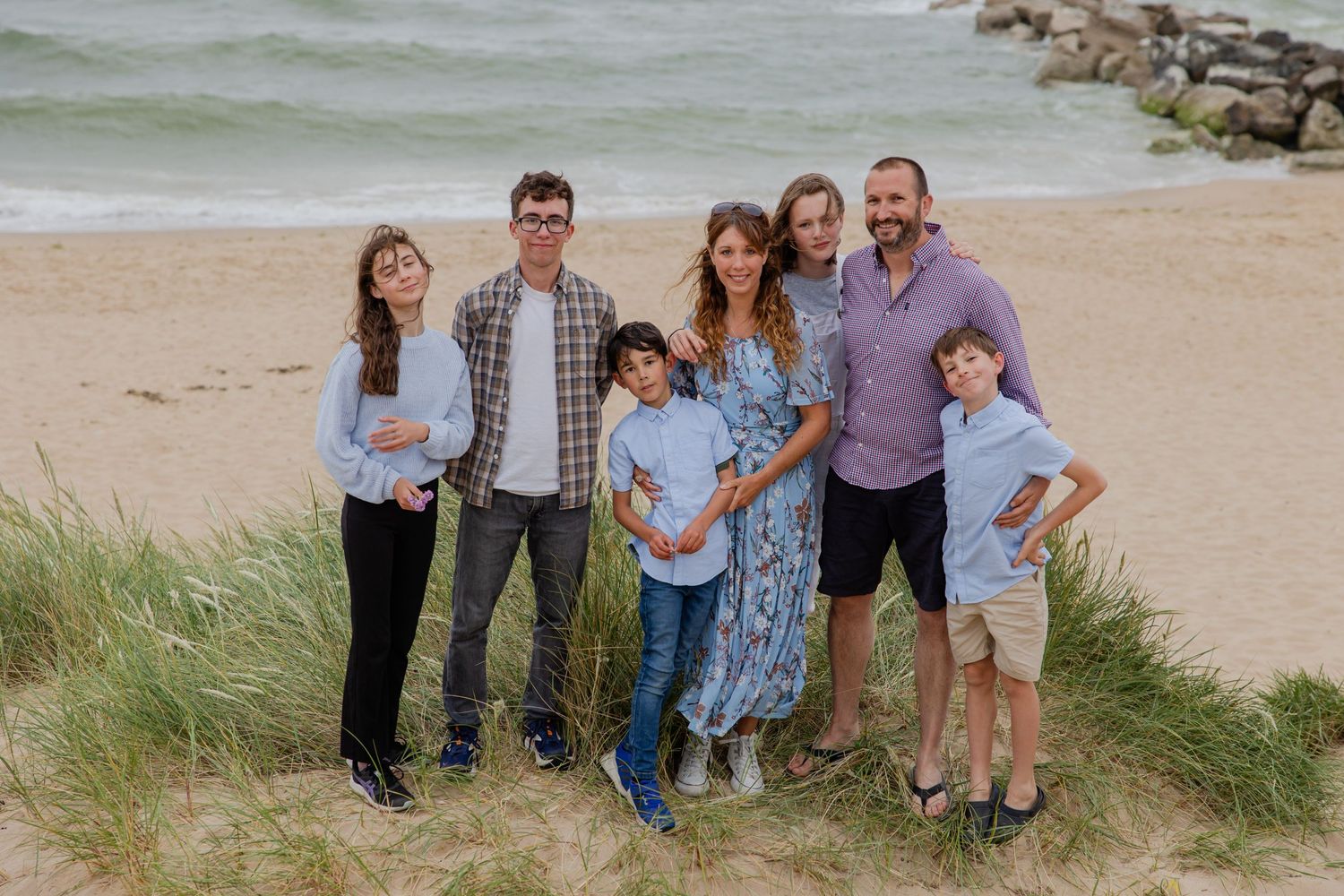 Family standing together on beach dunes with ocean and rocks visible in background.